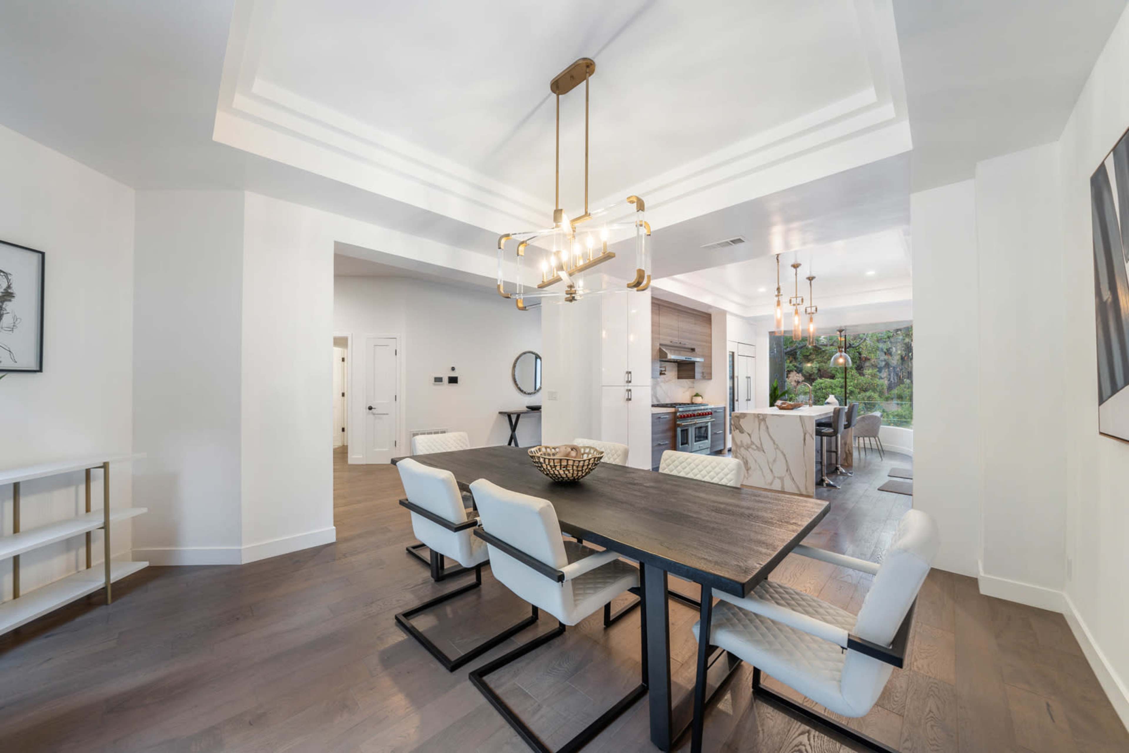A modern dining area with a rectangular table and white chairs, featuring a chandelier above and an open kitchen in the background.