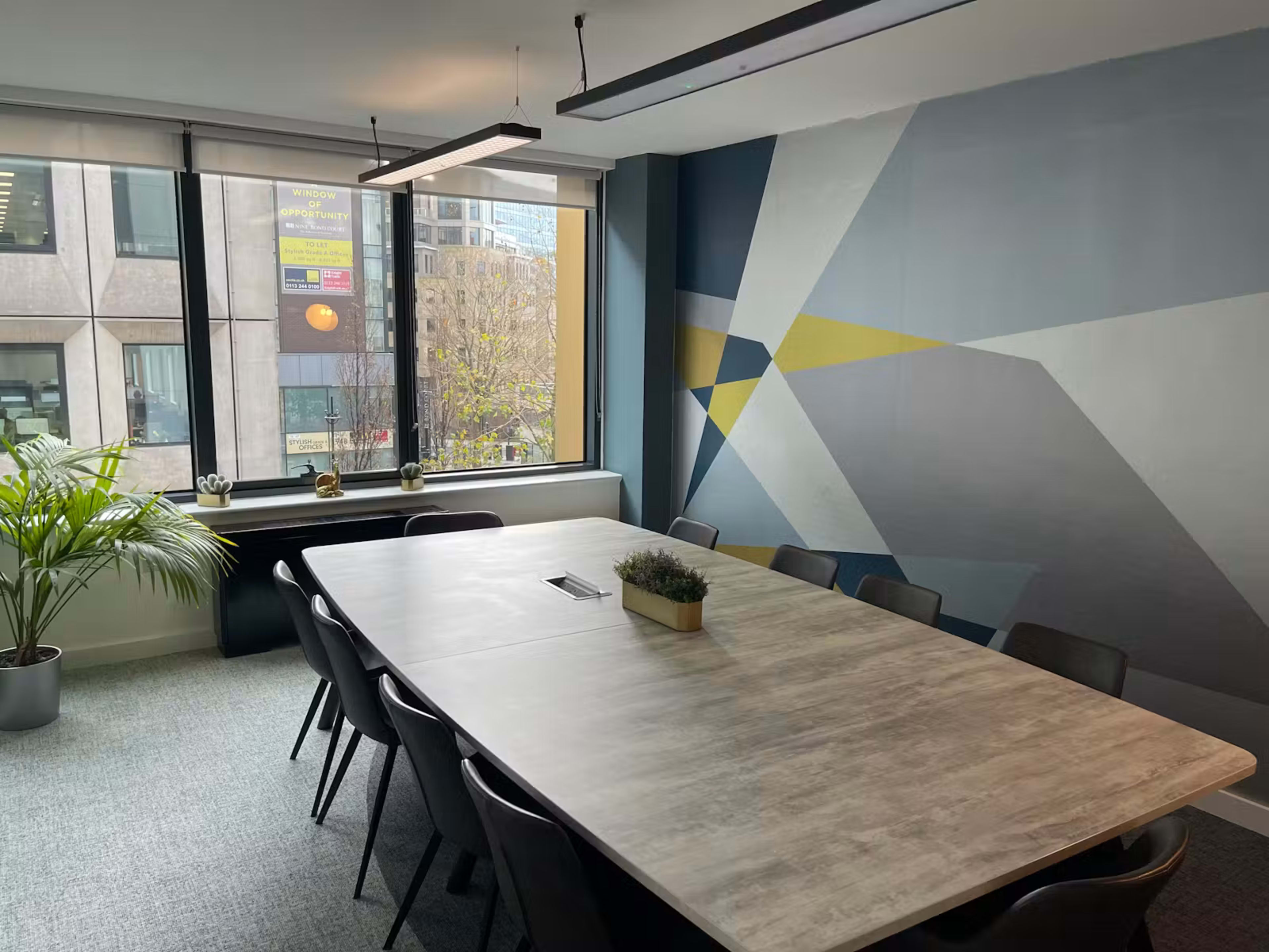 The image shows a modern conference room featuring a large wooden table surrounded by black chairs, with a vibrant geometric mural on one wall and views of a city through large windows.