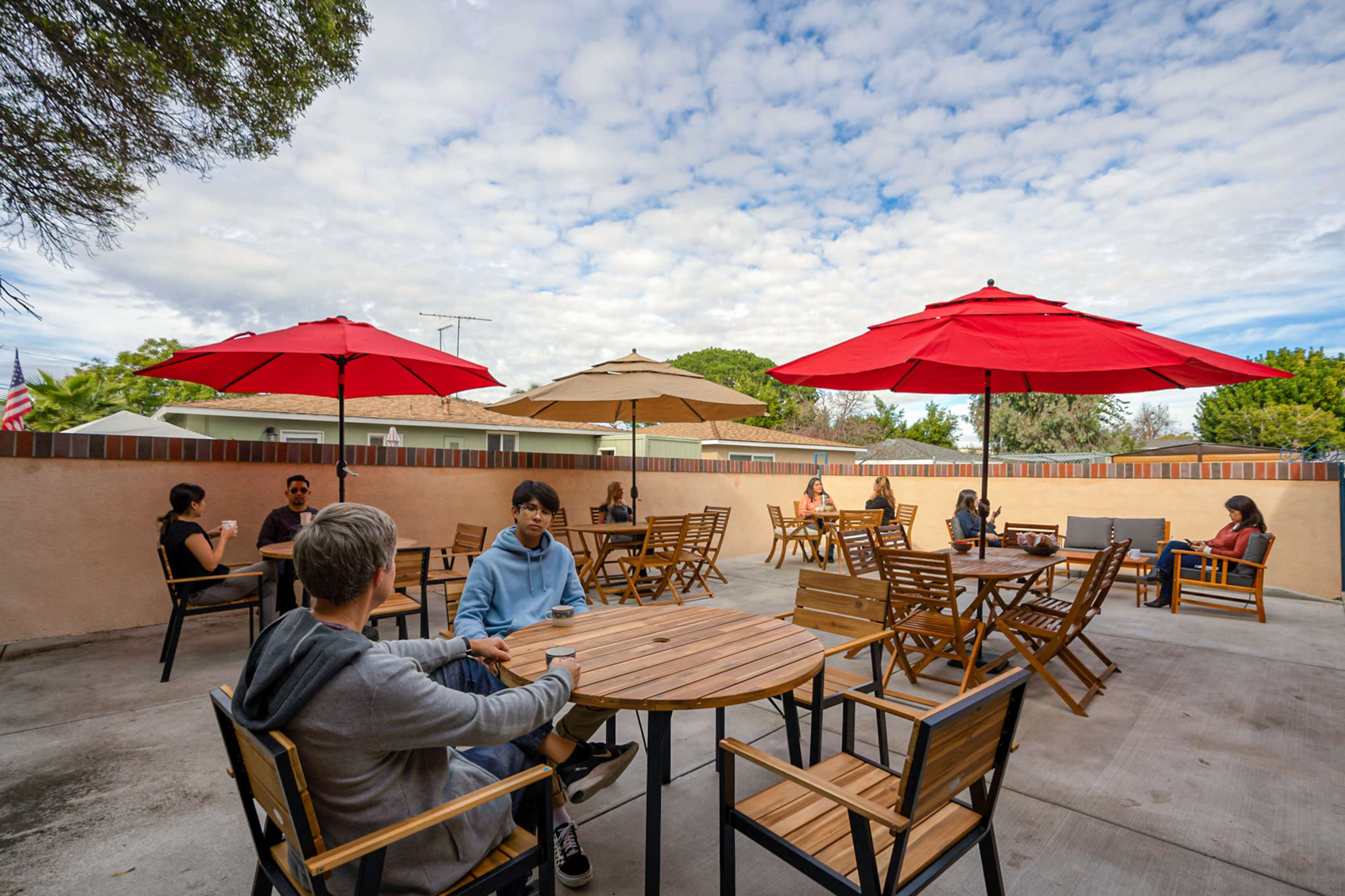 A group of people sit at wooden tables under various umbrellas in an outdoor seating area surrounded by a low wall and a cloudy sky.