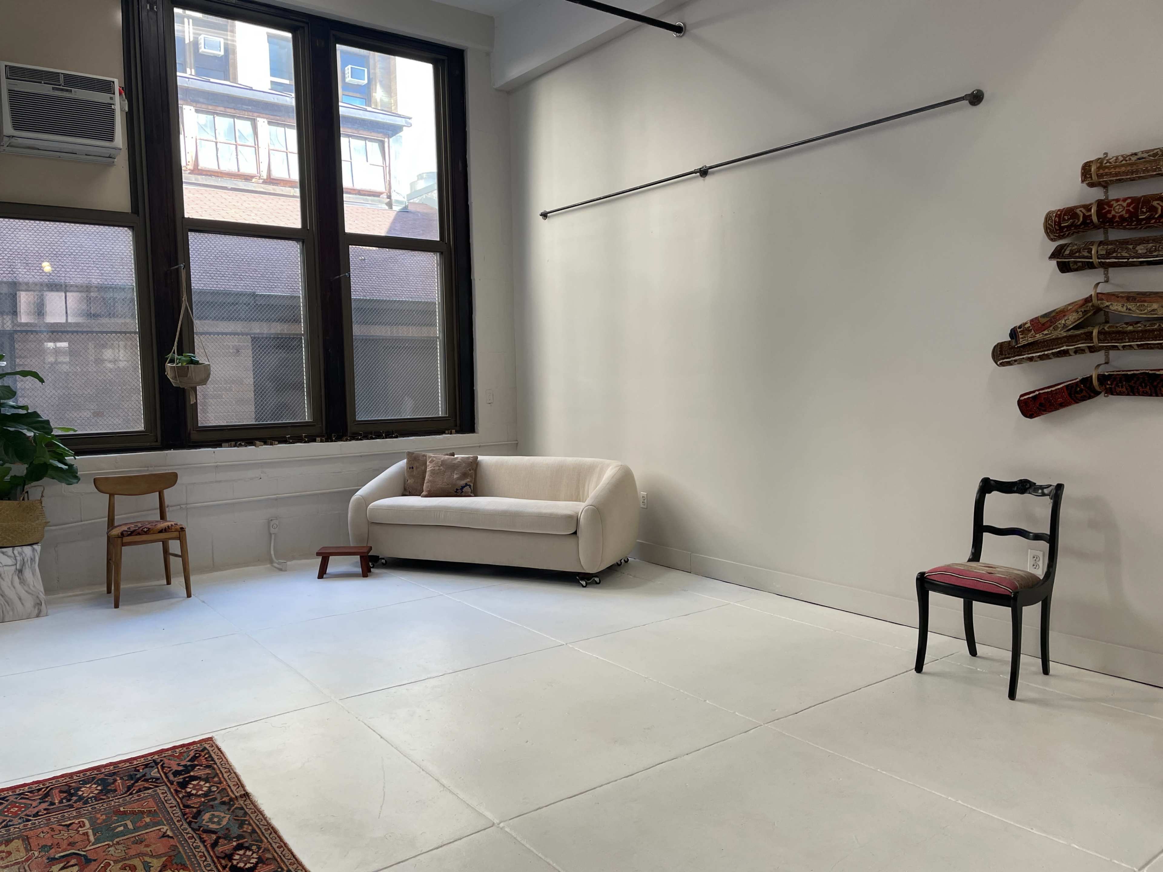 A minimalist indoor space with large windows, a beige couch, a black chair, and a patterned rug on the white floor.