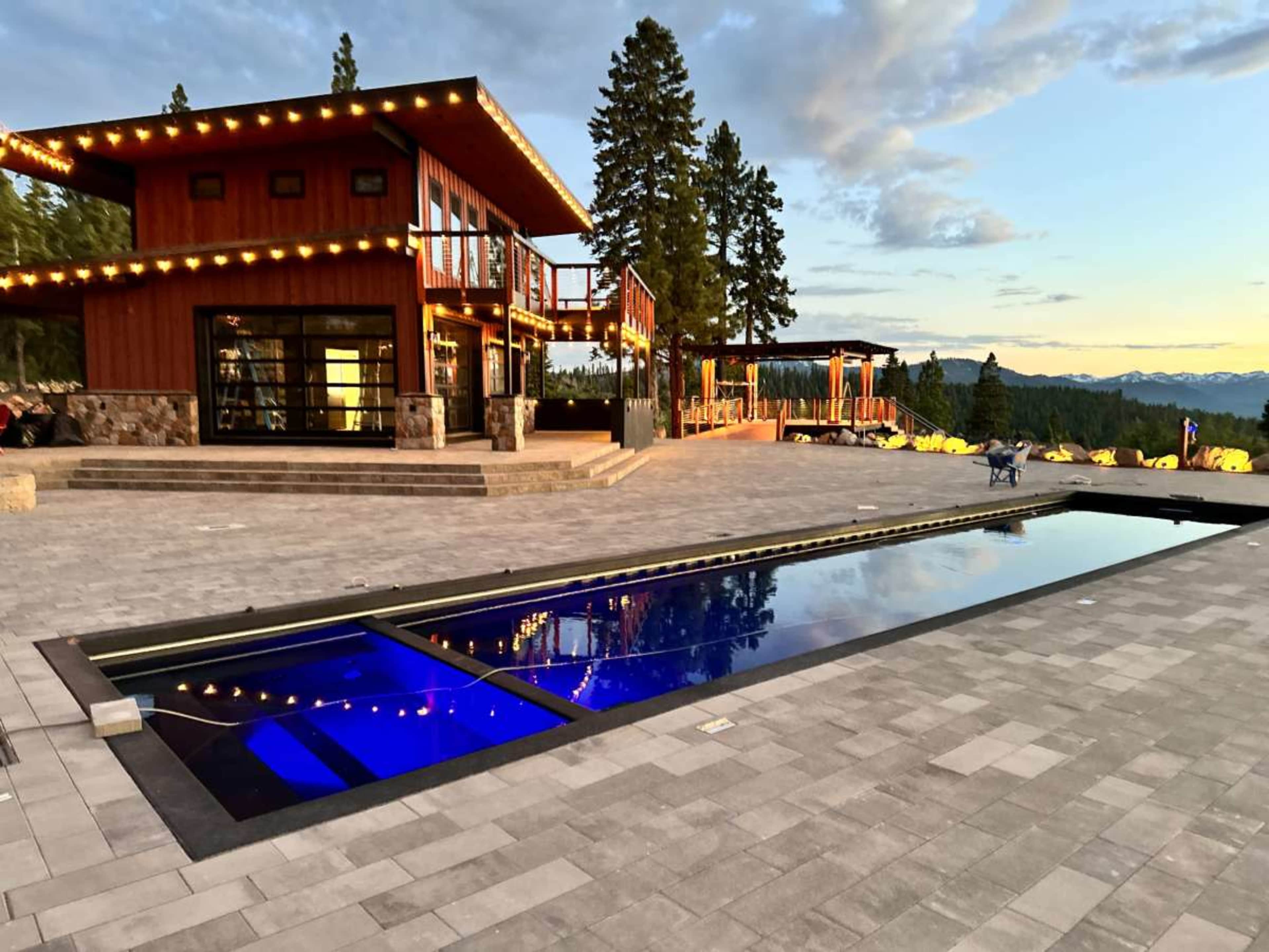 A modern, two-story house with large windows and outdoor lighting is situated next to a rectangular swimming pool on a stone patio overlooking a mountainous landscape.