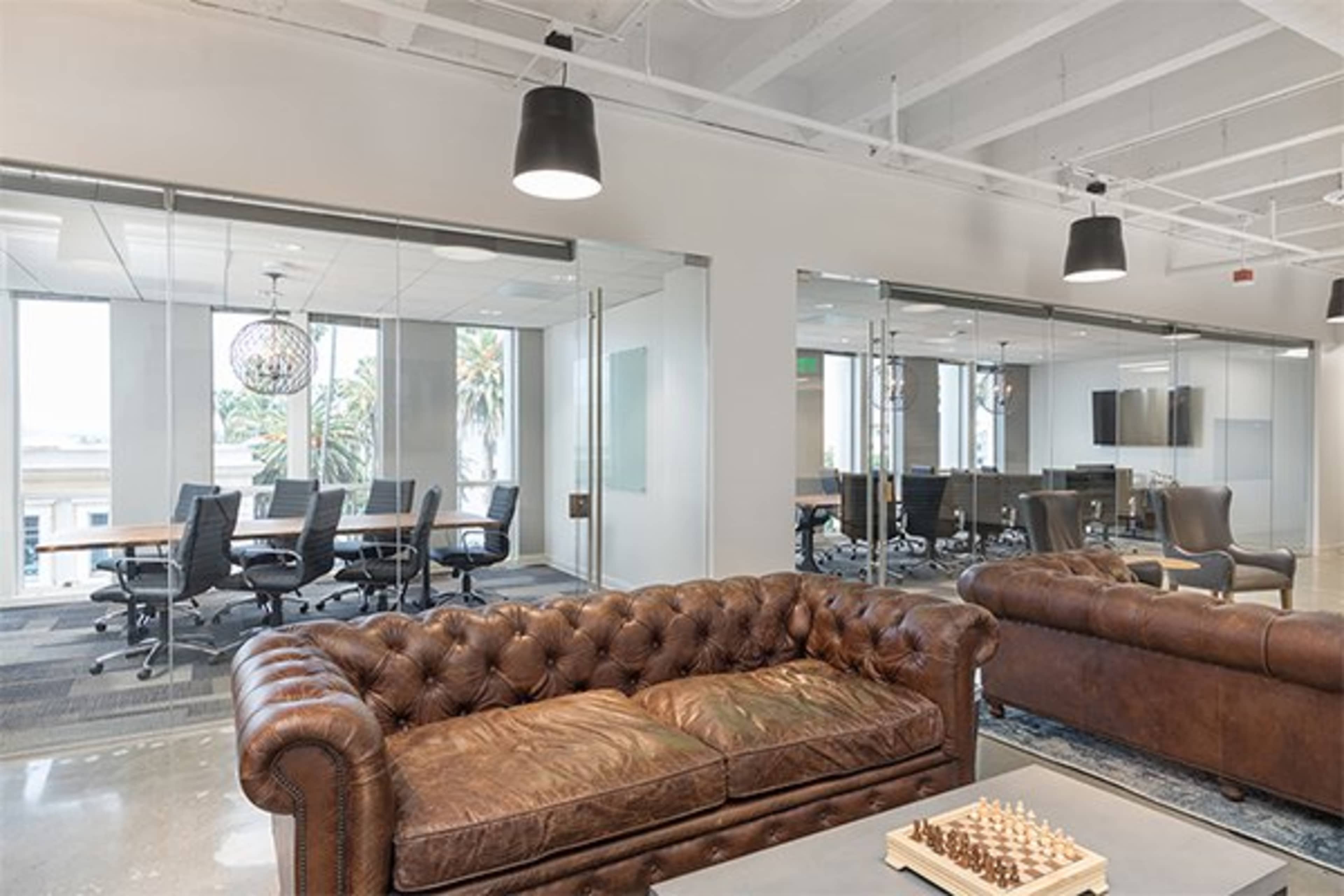 A modern office space featuring a brown leather sofa, glass conference rooms with large tables, and large windows allowing natural light in.