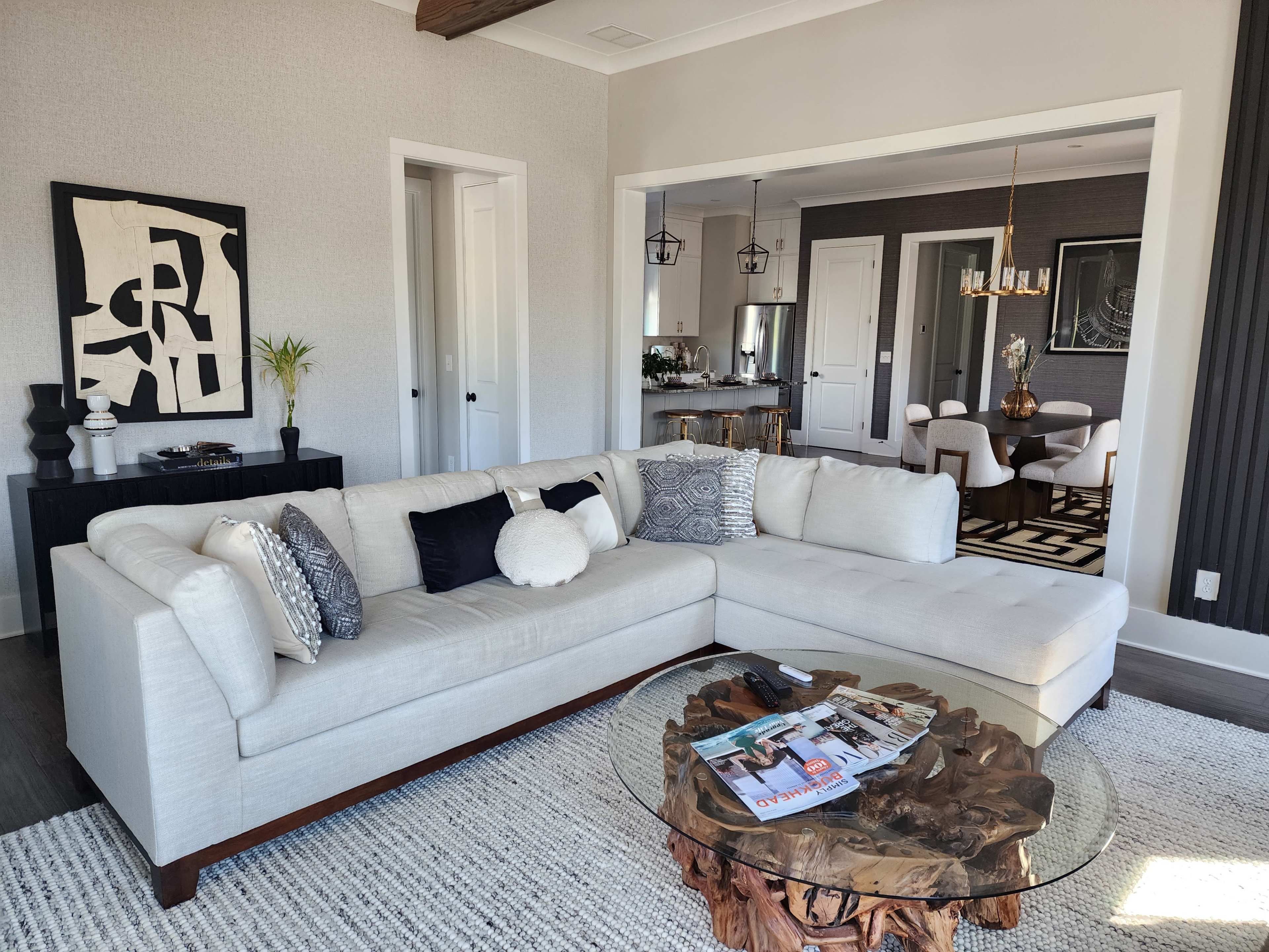 A light-colored sectional sofa is positioned in a modern living room, adjacent to a dining area featuring a dark color scheme, with a decorative coffee table made of wood and glass in front.