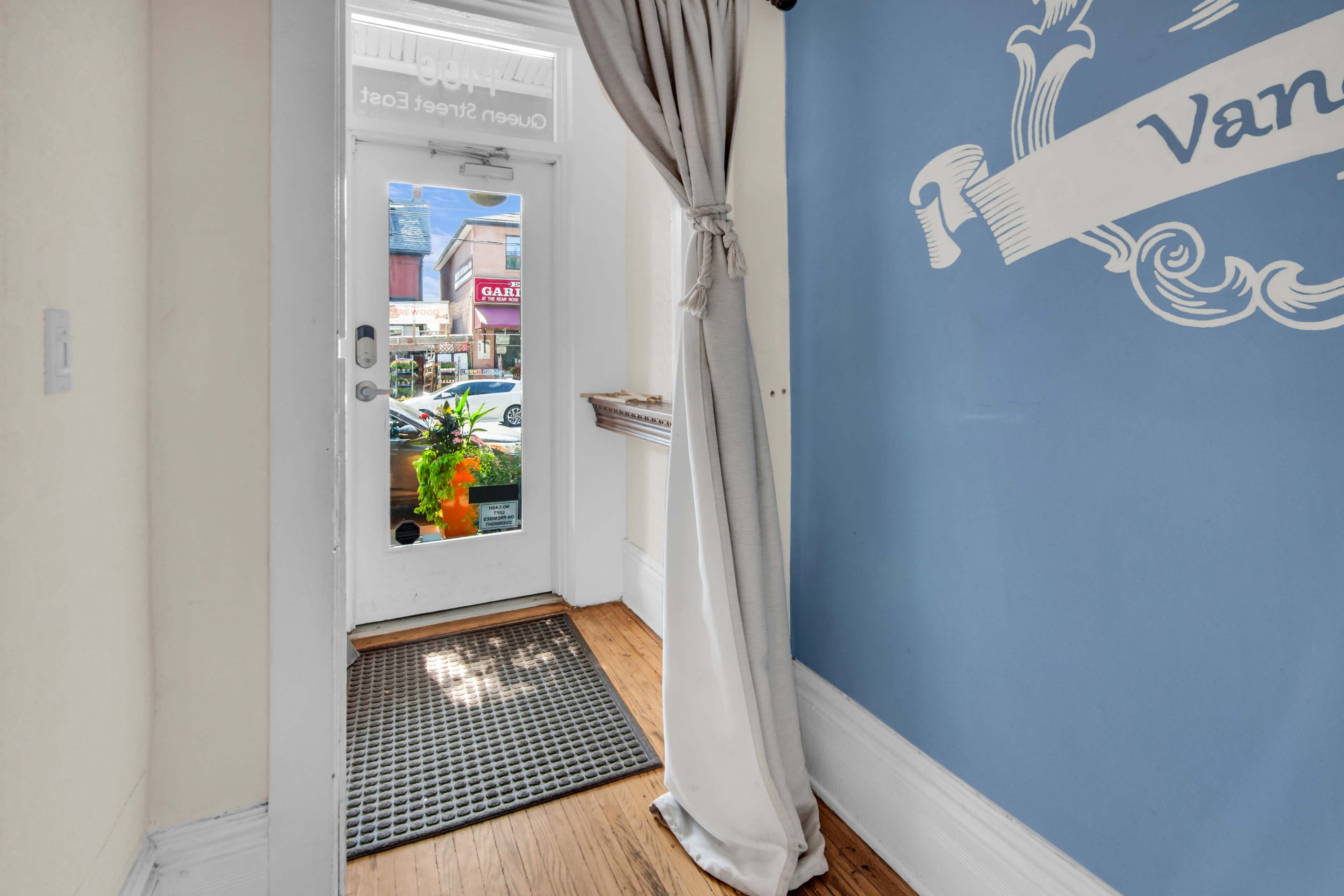 The image shows an entrance hall with a blue wall, a curtain to the side, a door leading outside, and a doormat on the wooden floor.