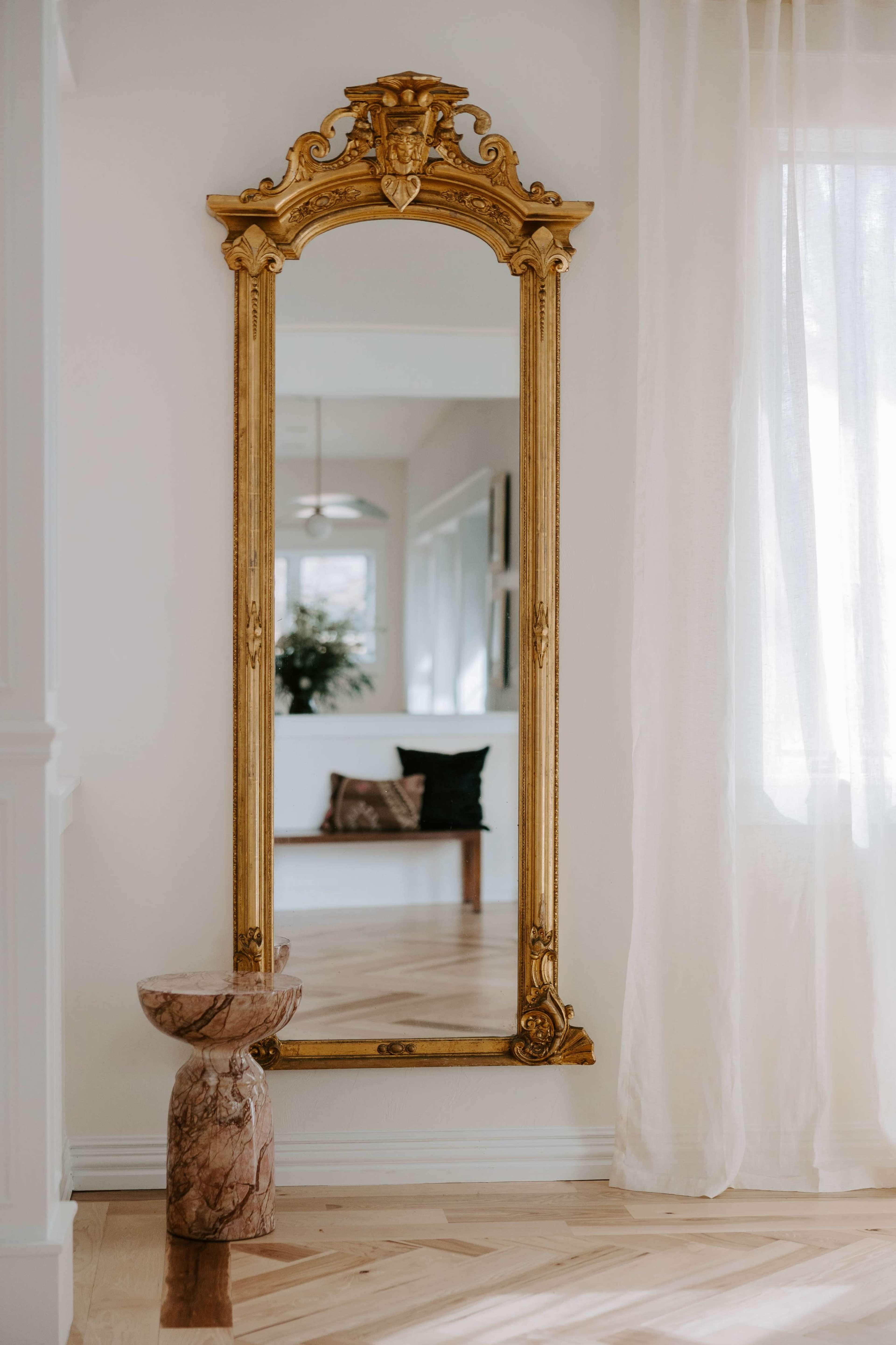 A large, ornate gold-framed mirror stands against a light-colored wall next to a round wooden side table.
