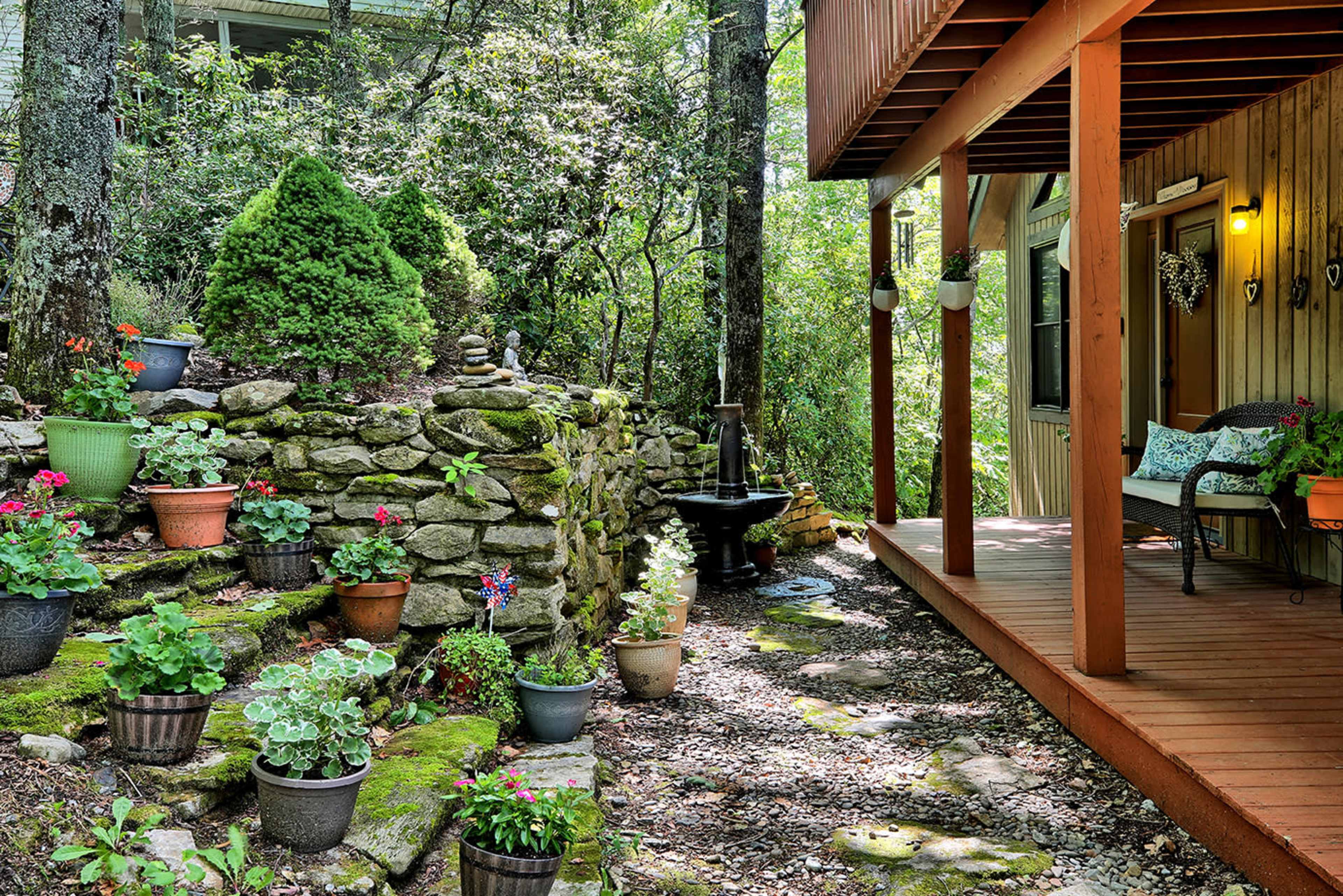 A stone wall garden path with various potted plants leads to a wooden deck surrounded by trees.