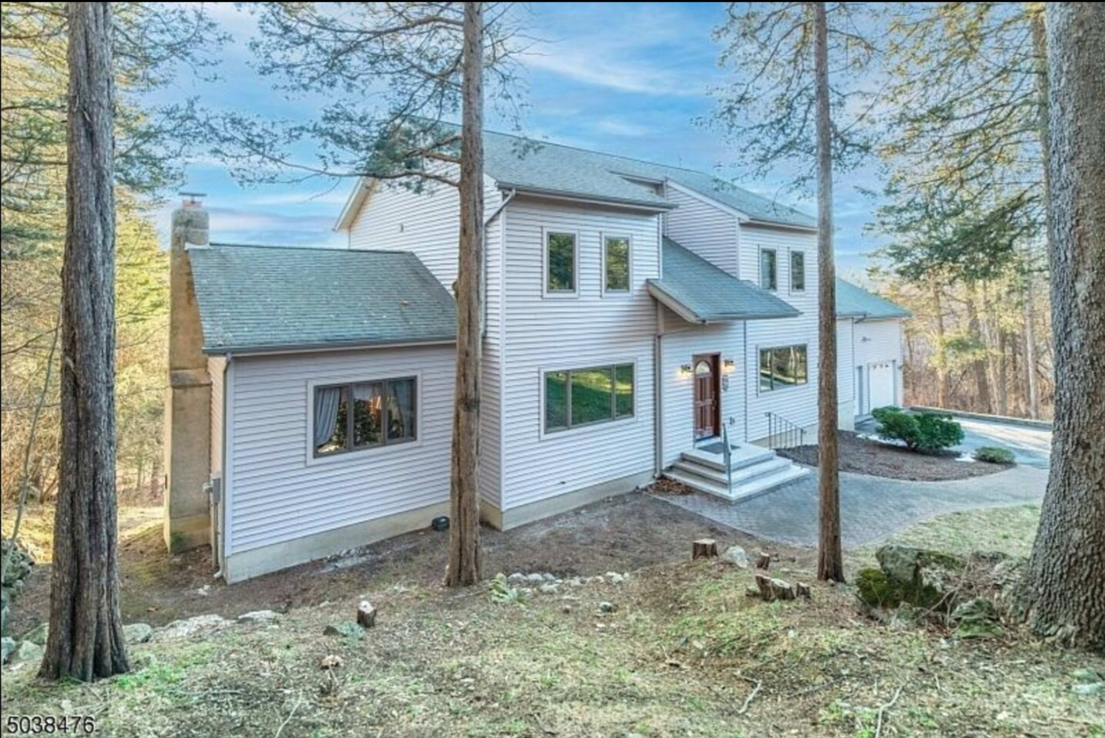 A two-story house with light-colored siding is situated among tall trees, featuring a front porch and a paved driveway.
