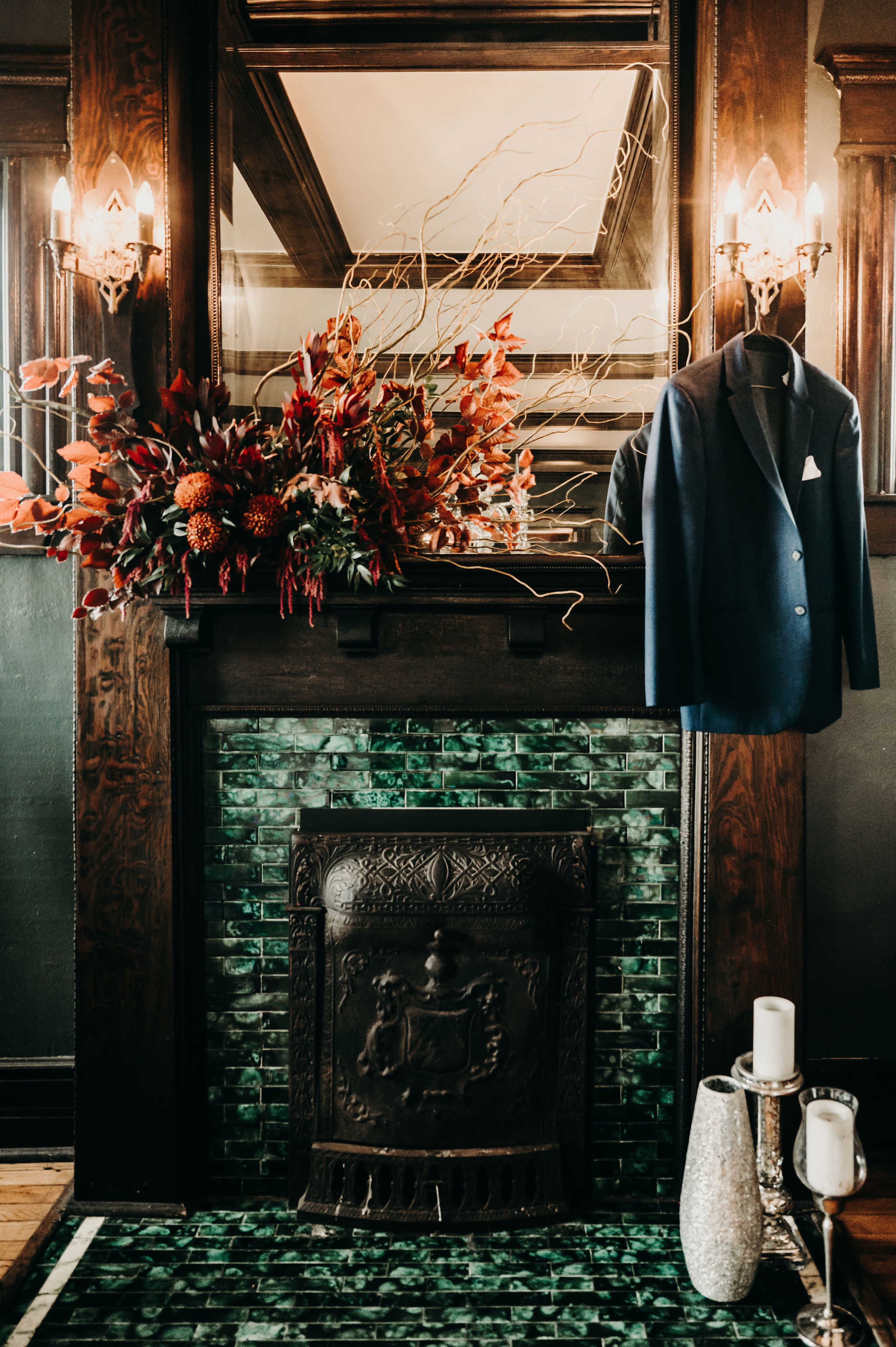 A dark wooden mantel displays an arrangement of autumnal flowers above a vintage green-tiled fireplace, with a navy suit jacket hanging nearby.