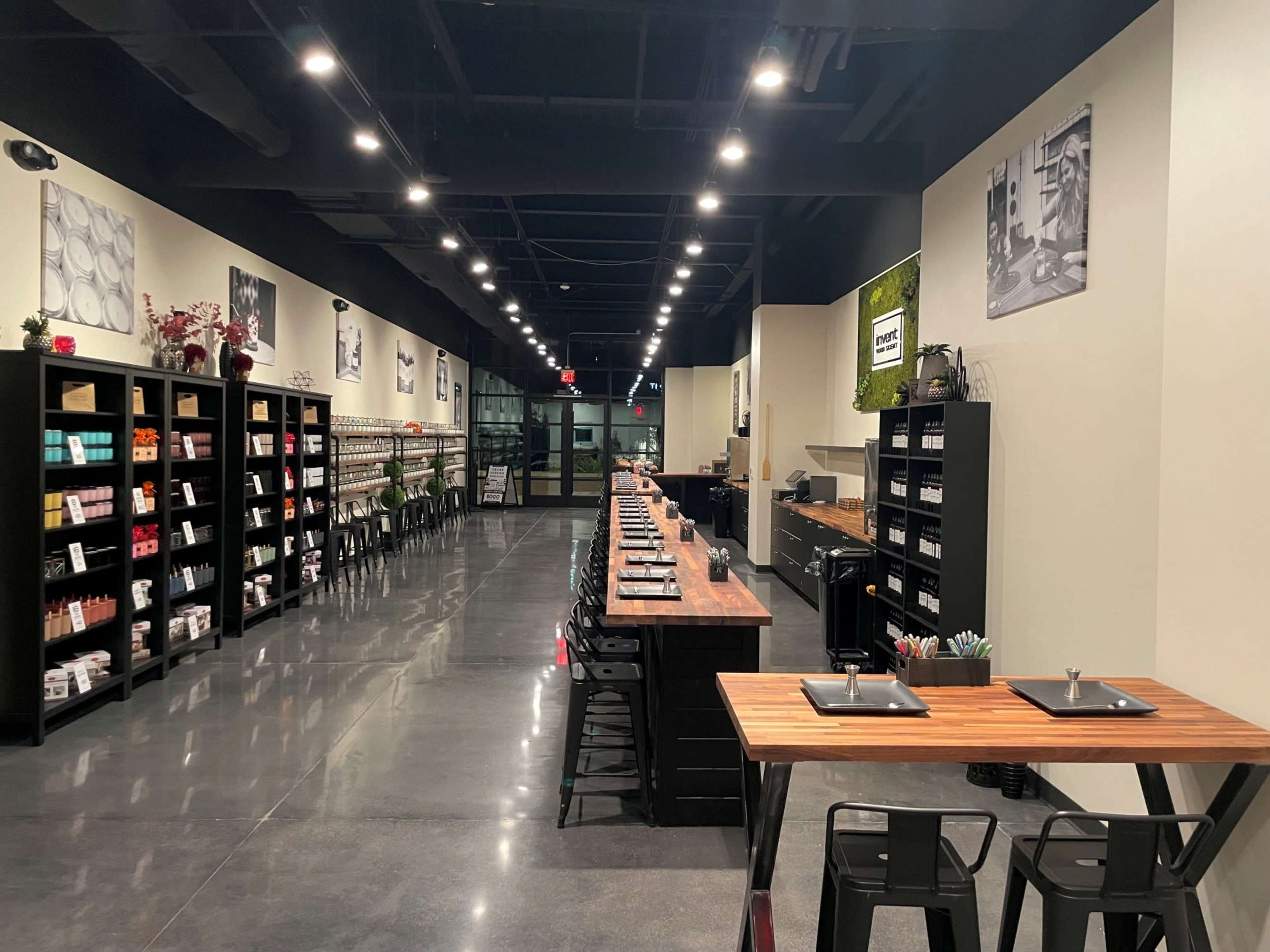 The image shows a modern interior of a well-lit retail space featuring shelves filled with various products and a long communal table set for several guests.