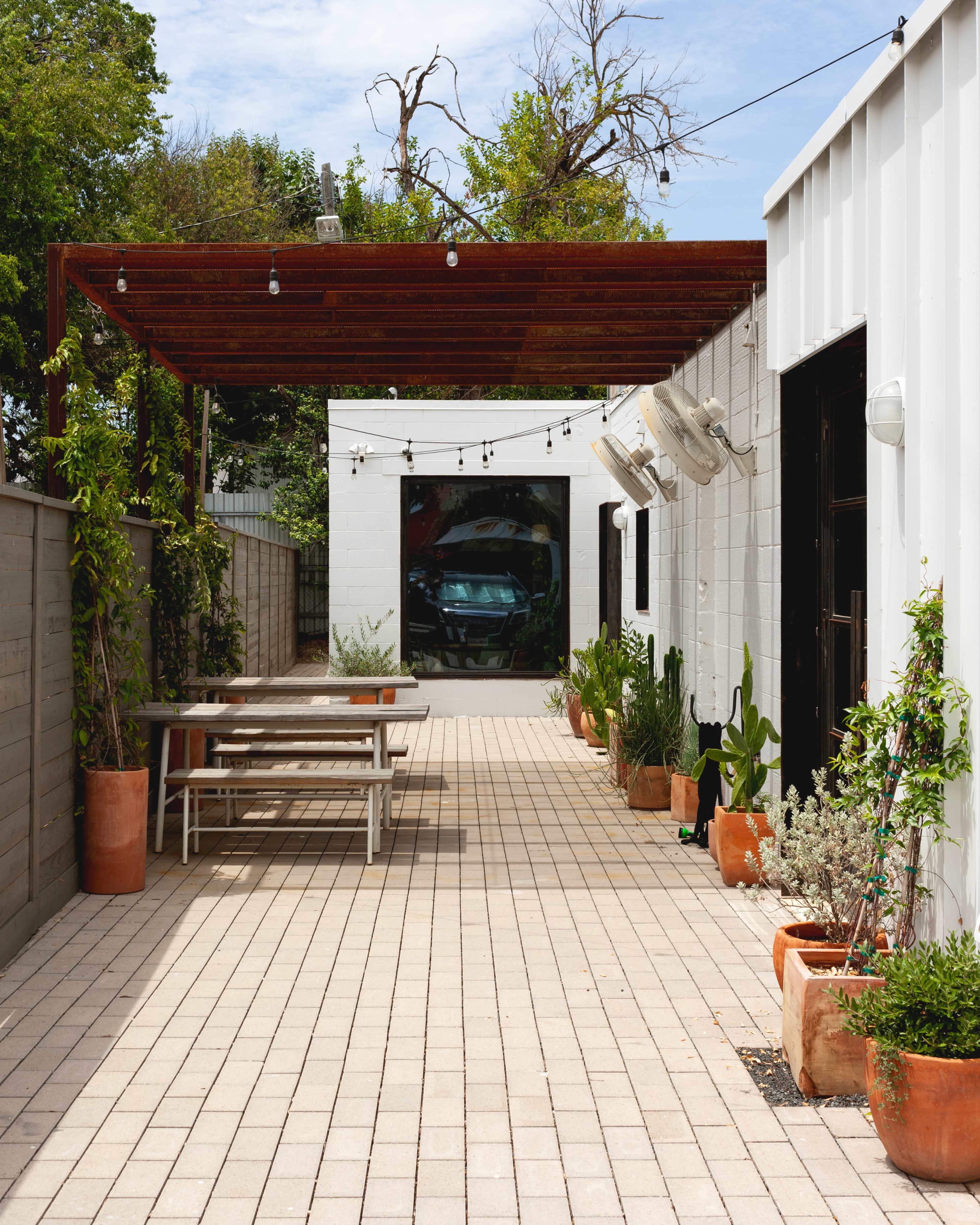 The image shows a paved outdoor area with a table, various potted plants, and a car visible in a garage at the end of the space, all under a shaded wooden structure.