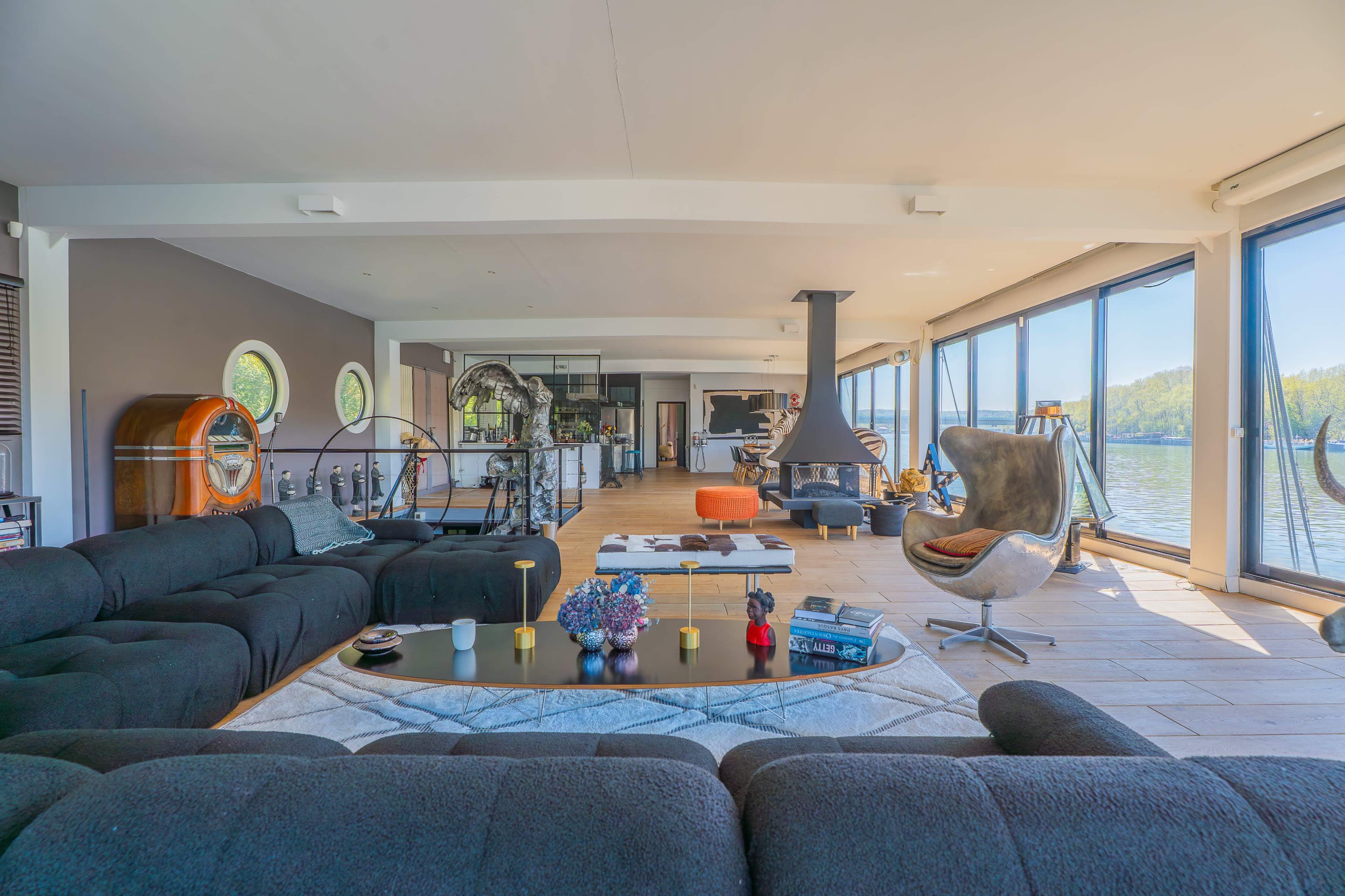 The image depicts a modern, open-concept living space with large windows overlooking a body of water, featuring sleek furnishings and a minimalist design.