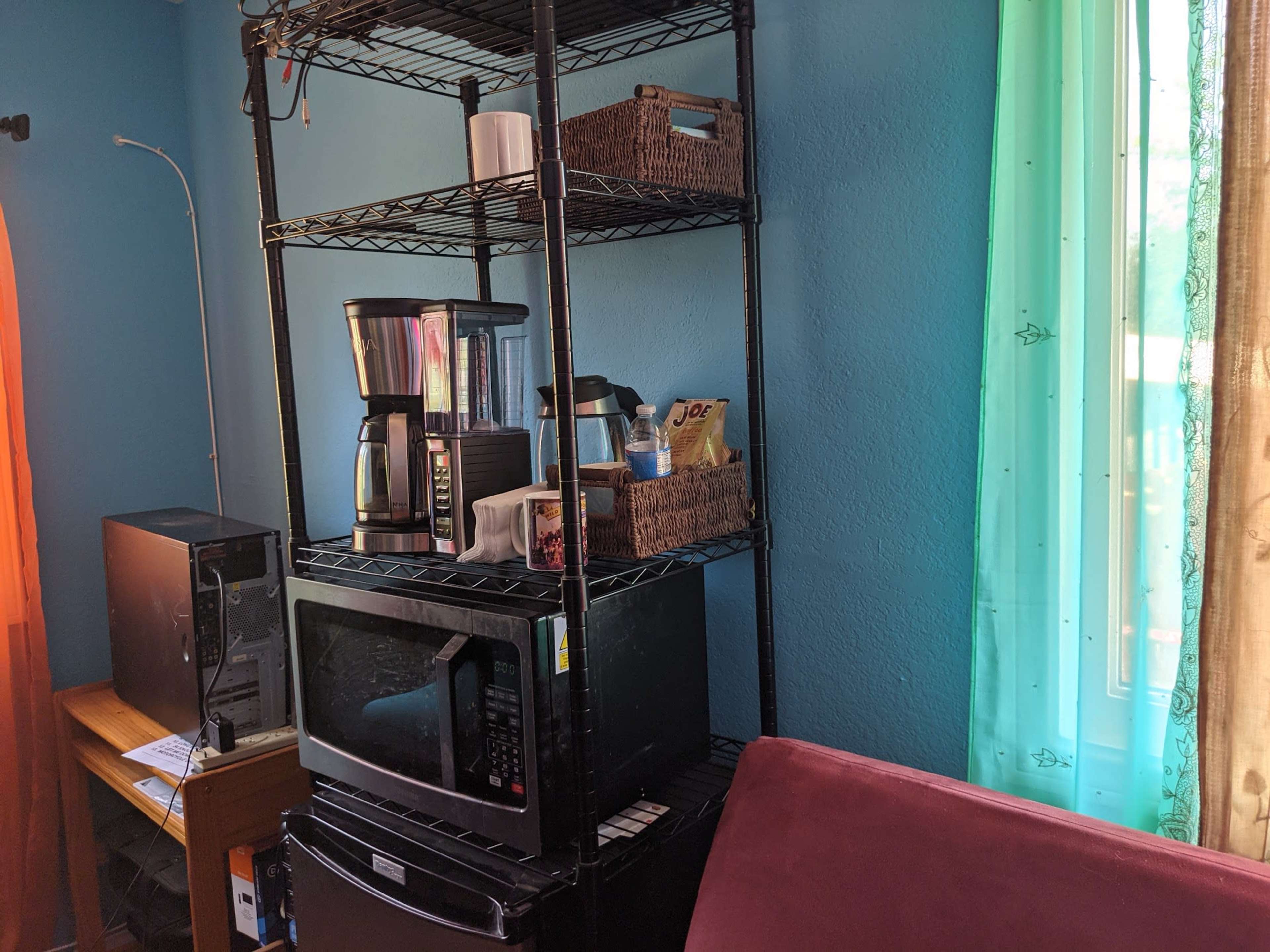 A kitchen corner with a black shelving unit that holds a coffee maker, microwave, and various baskets, alongside a window with sheer curtains.
