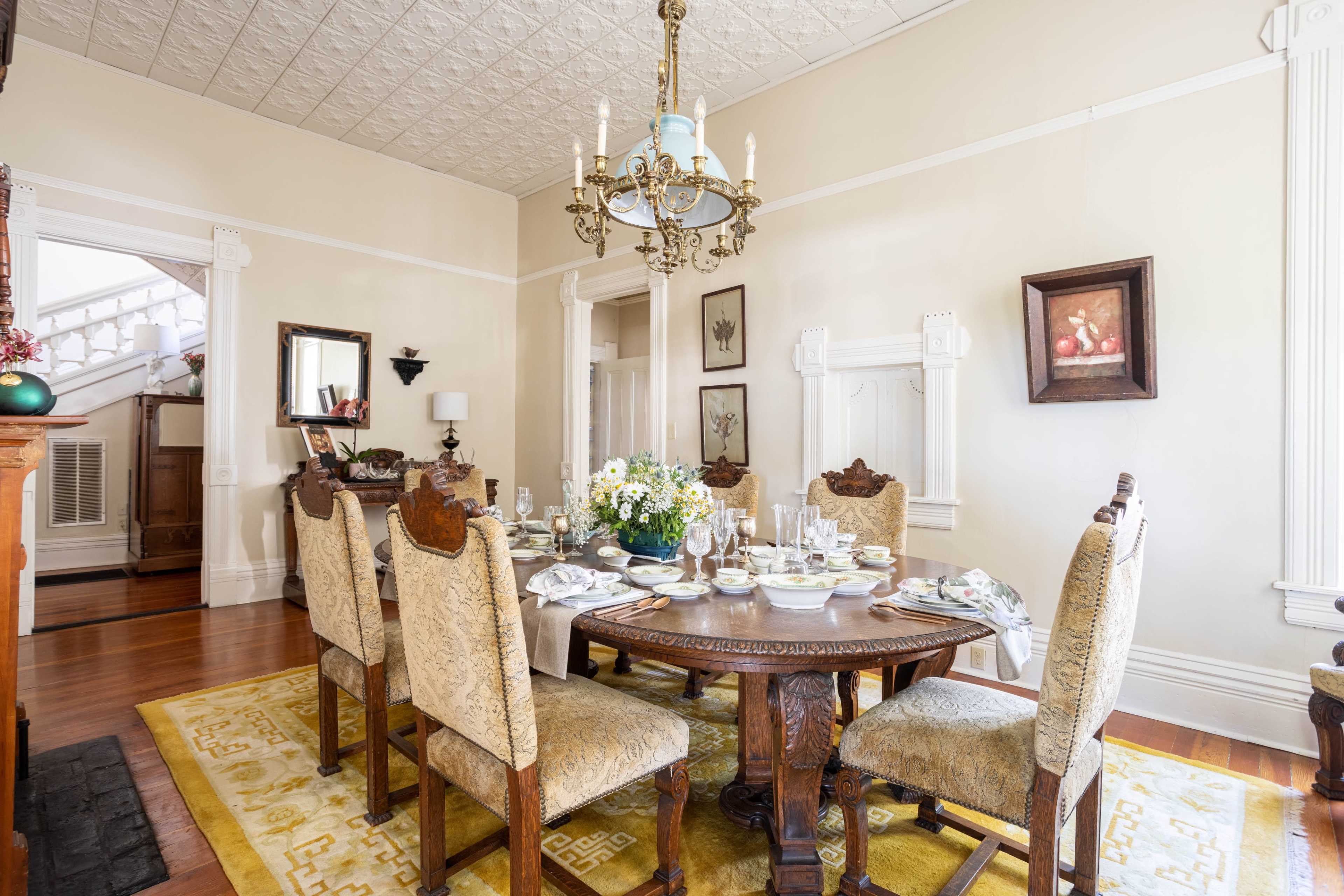 A formal dining room features a large wooden table set for a meal, surrounded by eight upholstered chairs and illuminated by an ornate chandelier.
