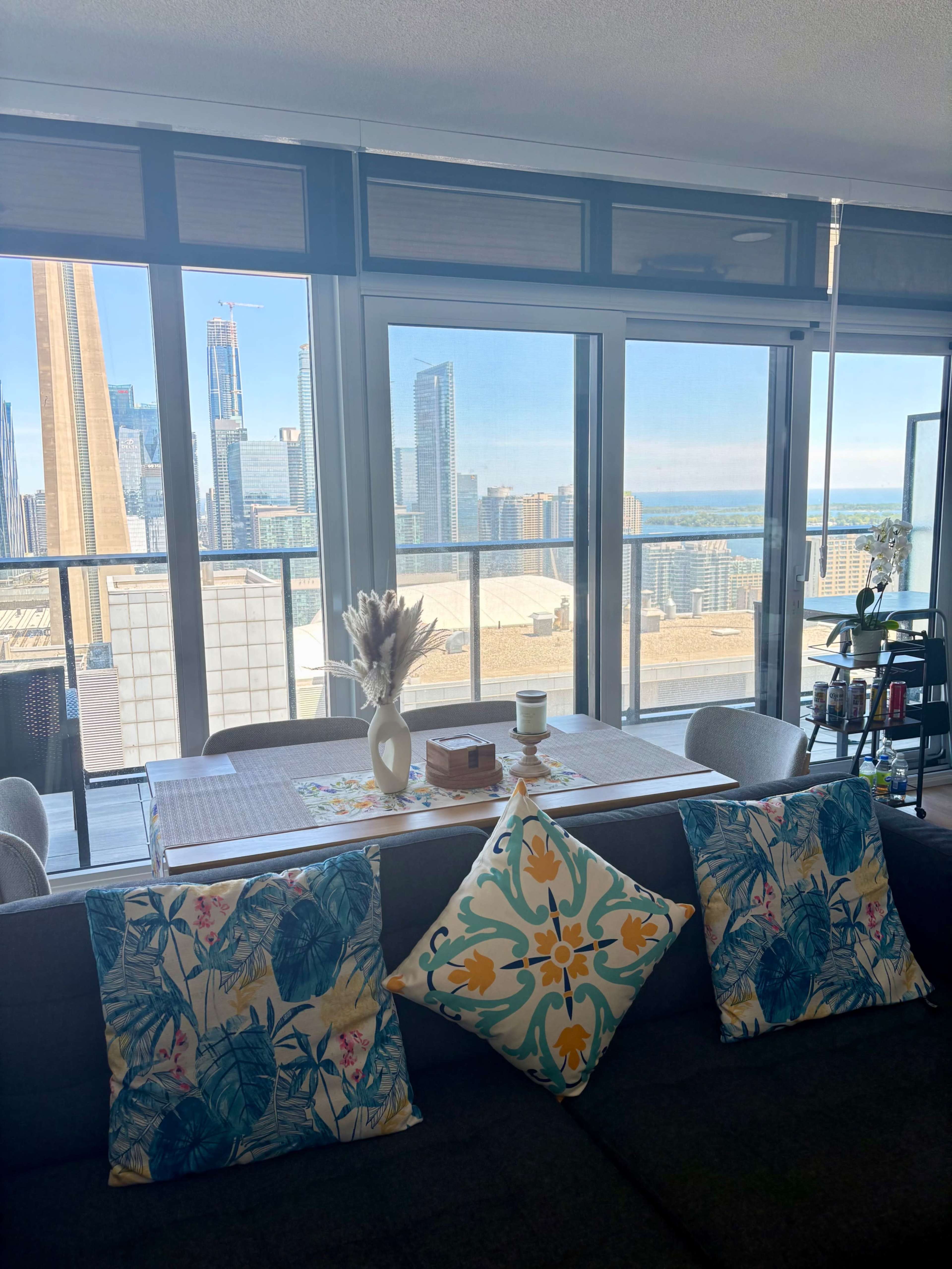 A modern apartment features a dining table with a scenic view of a city skyline and water, flanked by decorative pillows on a gray sofa.