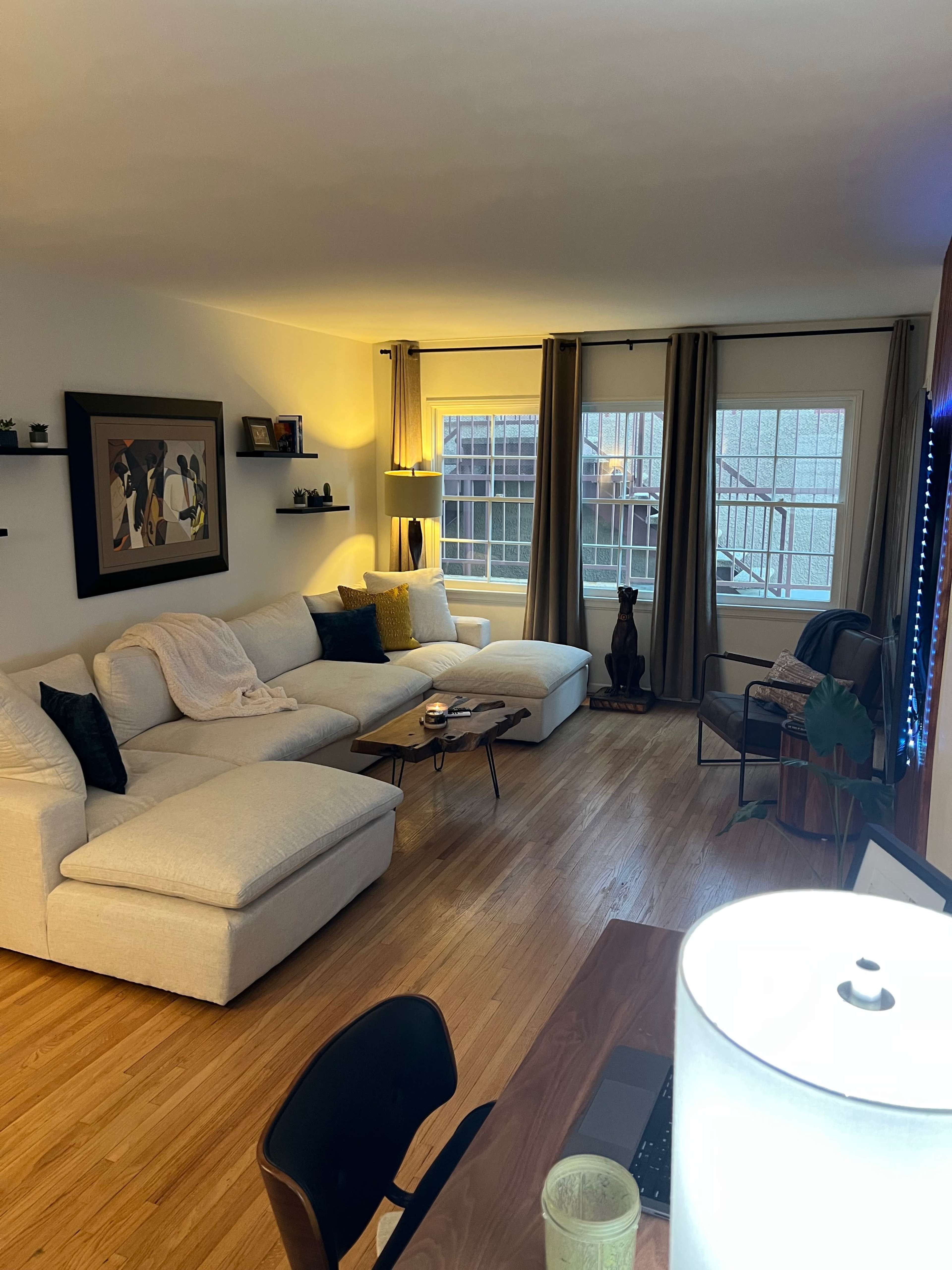 A bright living room features a large sectional sofa, a coffee table, and a desk, with large windows dressed in curtains.