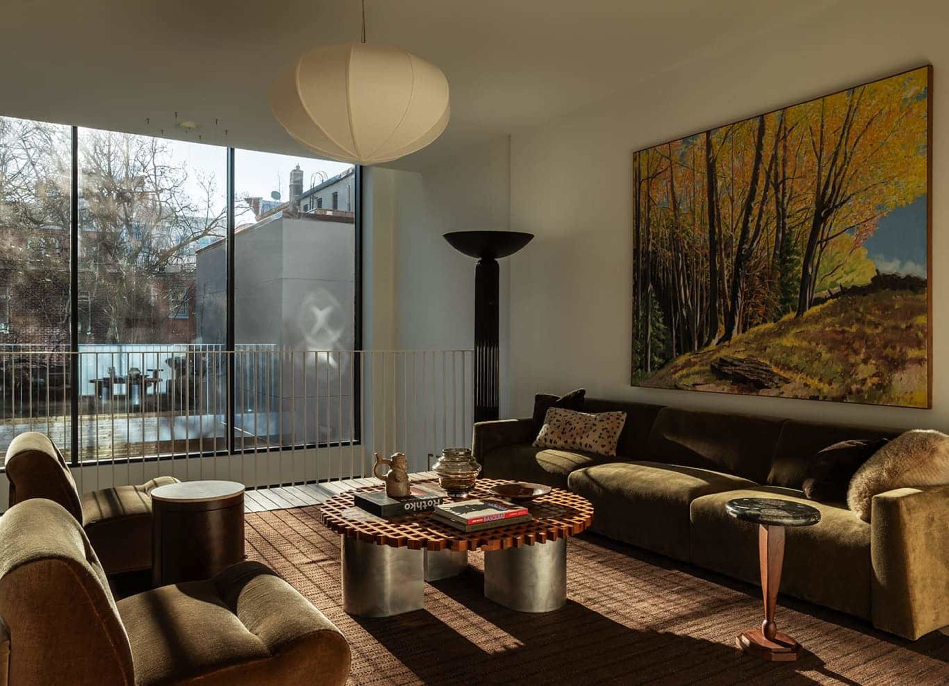 The image shows a modern living room with large windows, a round coffee table, a sectional sofa, and a colorful landscape painting on the wall.