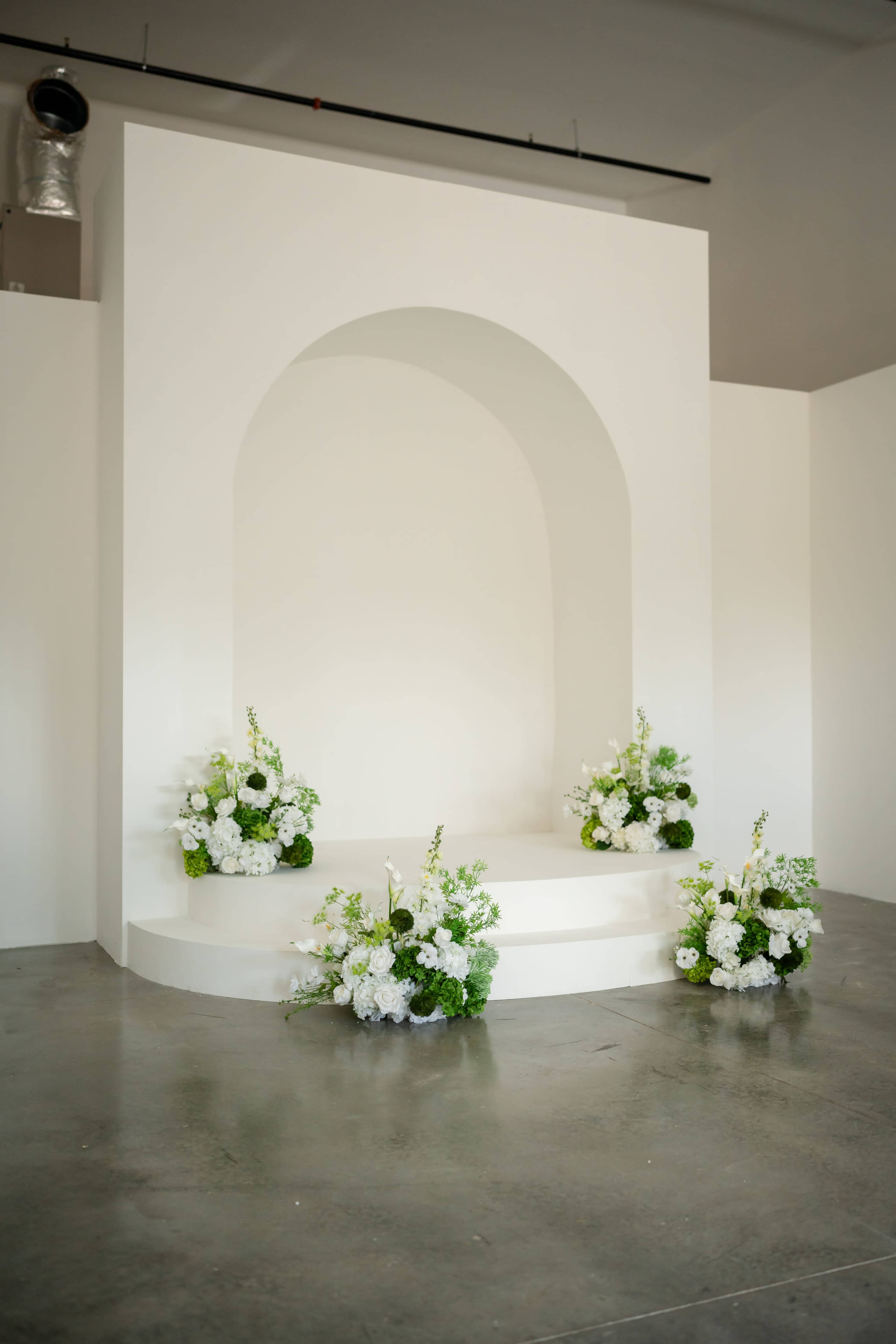 A minimalist white stage with an arched backdrop is adorned with floral arrangements on either side.