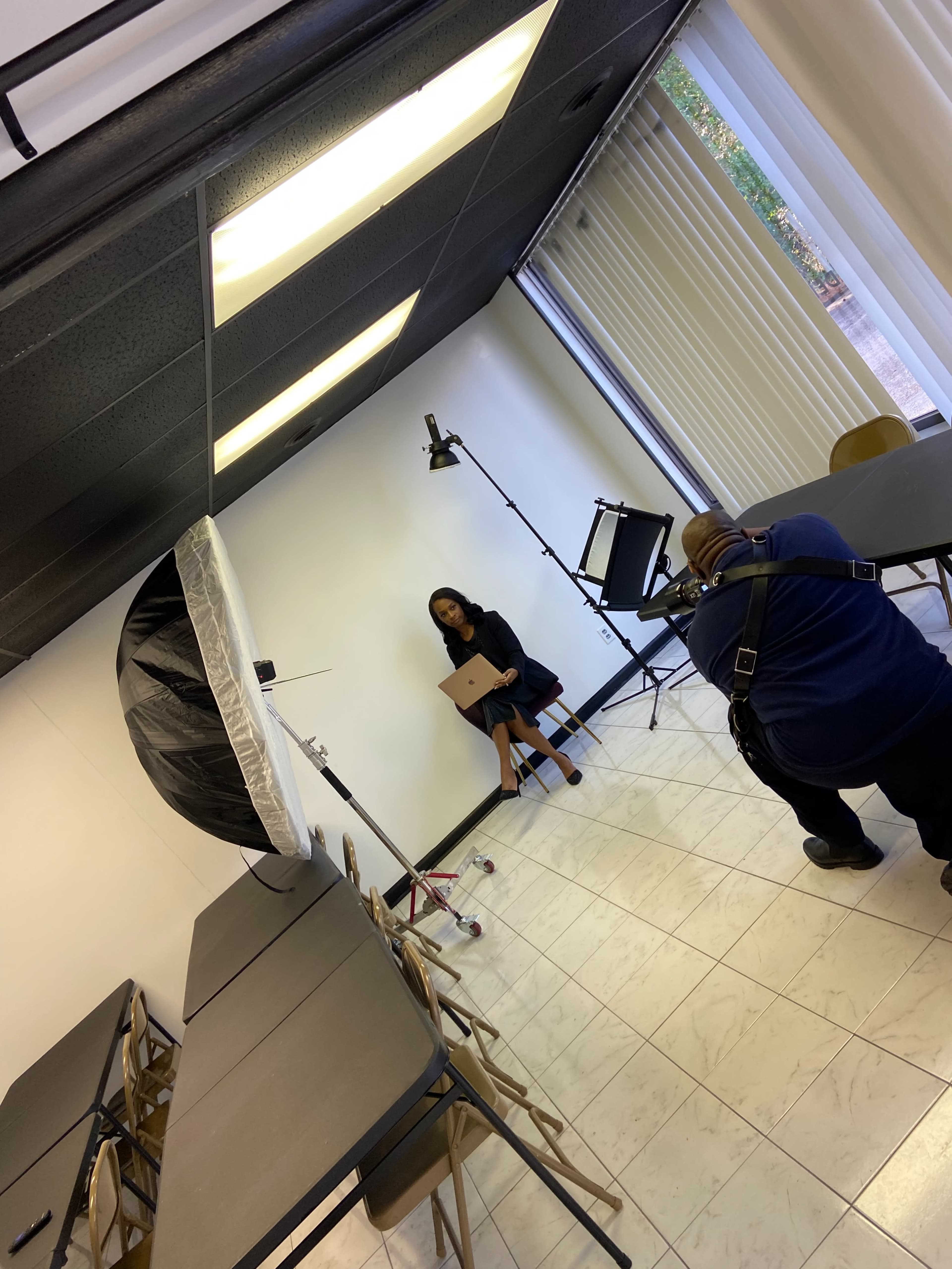 A photographer is capturing an image of a woman sitting with a laptop against a plain white wall in a studio setting.