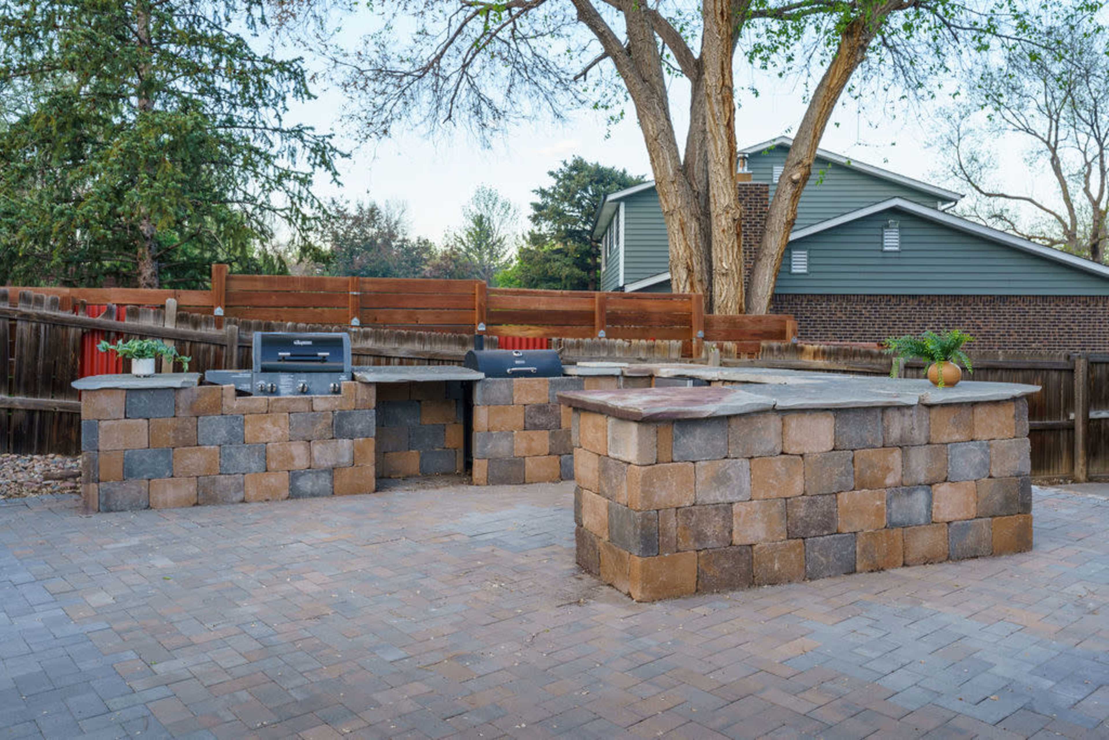 The image shows an outdoor kitchen area with stone countertops and built-in grills, surrounded by a paved patio and wooden fencing.