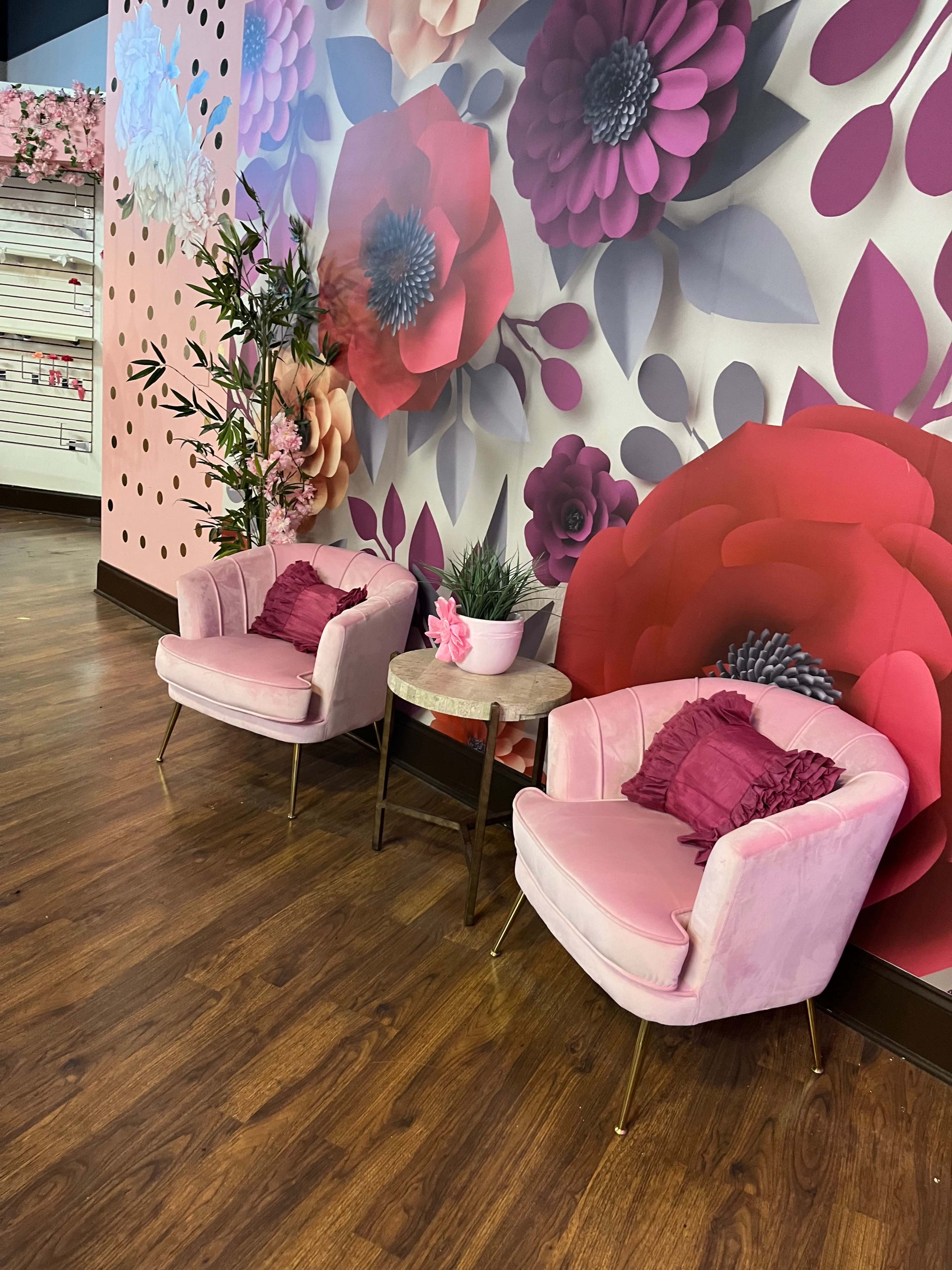 Two pink velvet chairs with decorative pillows are positioned next to a small round table against a backdrop of large floral wallpaper.
