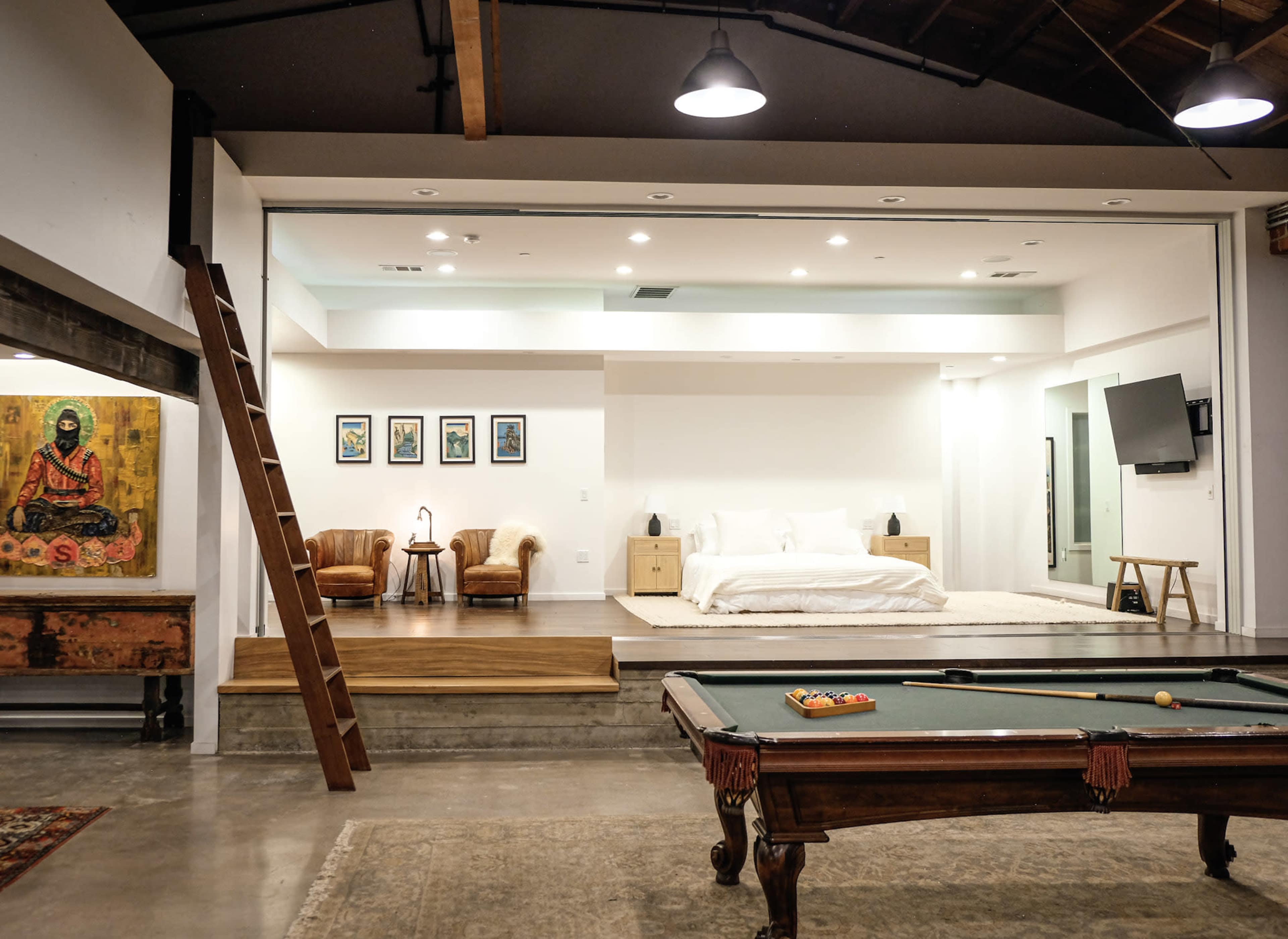 The image shows a spacious room featuring a pool table in the foreground, with a cozy sleeping area visible in the background.