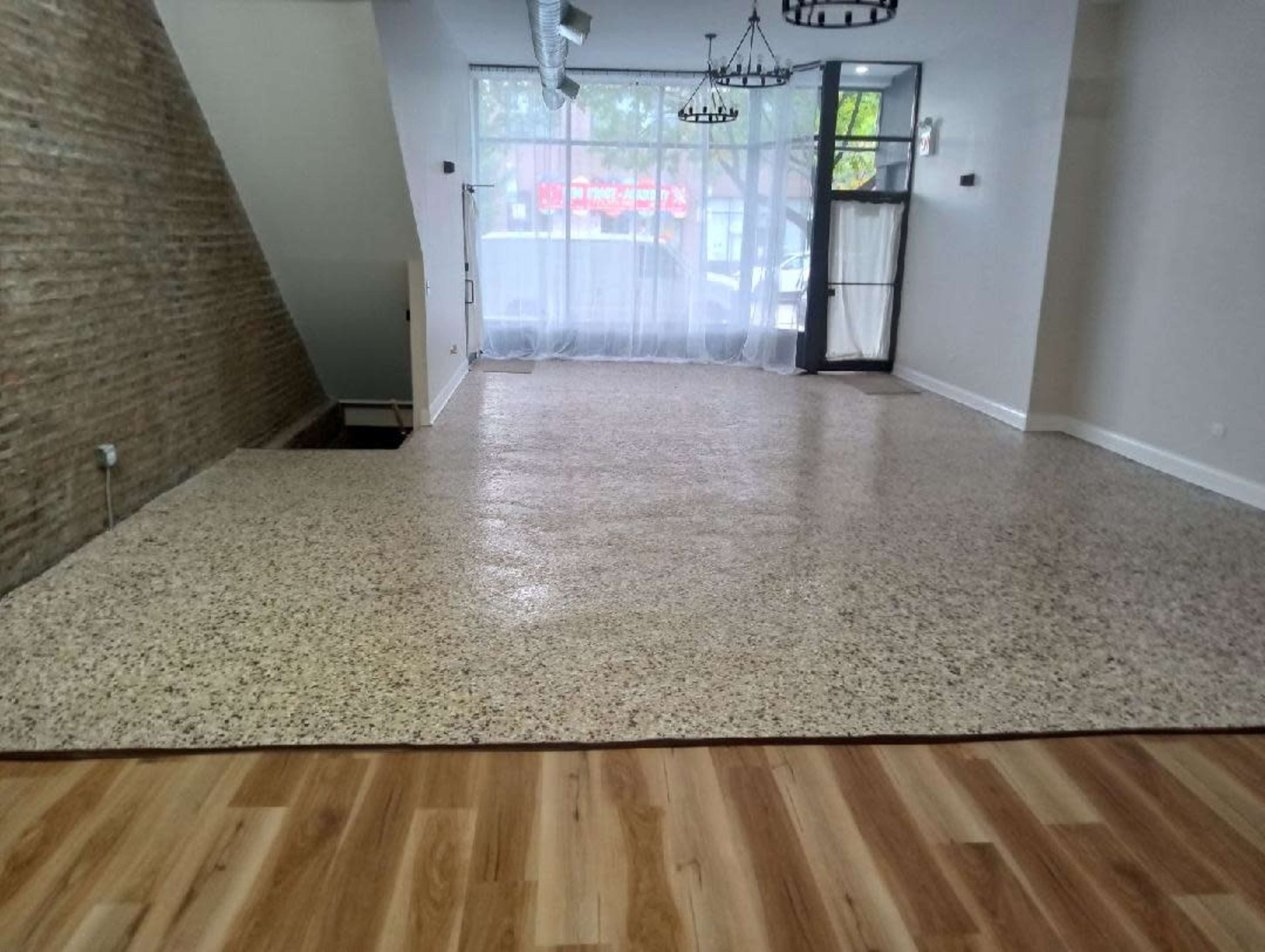 The image shows an interior space with a terrazzo floor and wooden flooring, featuring large windows that allow natural light to enter.