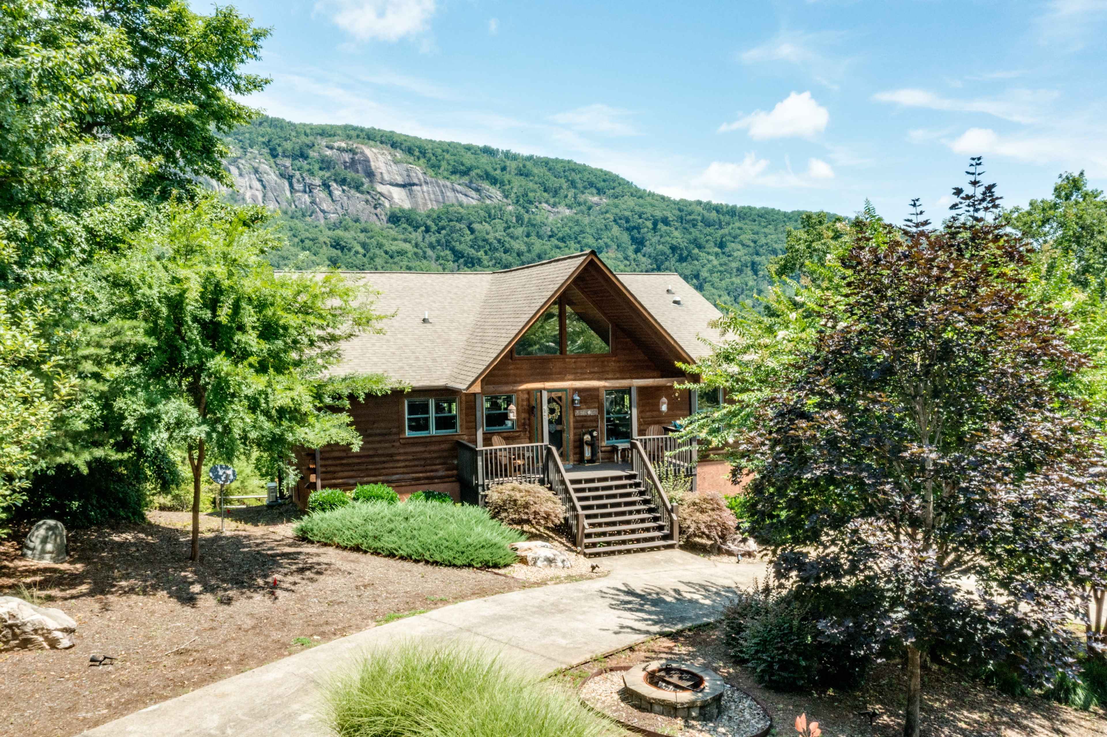 A wooden cabin with large windows and a front porch is situated in a landscaped area, backed by a mountain.