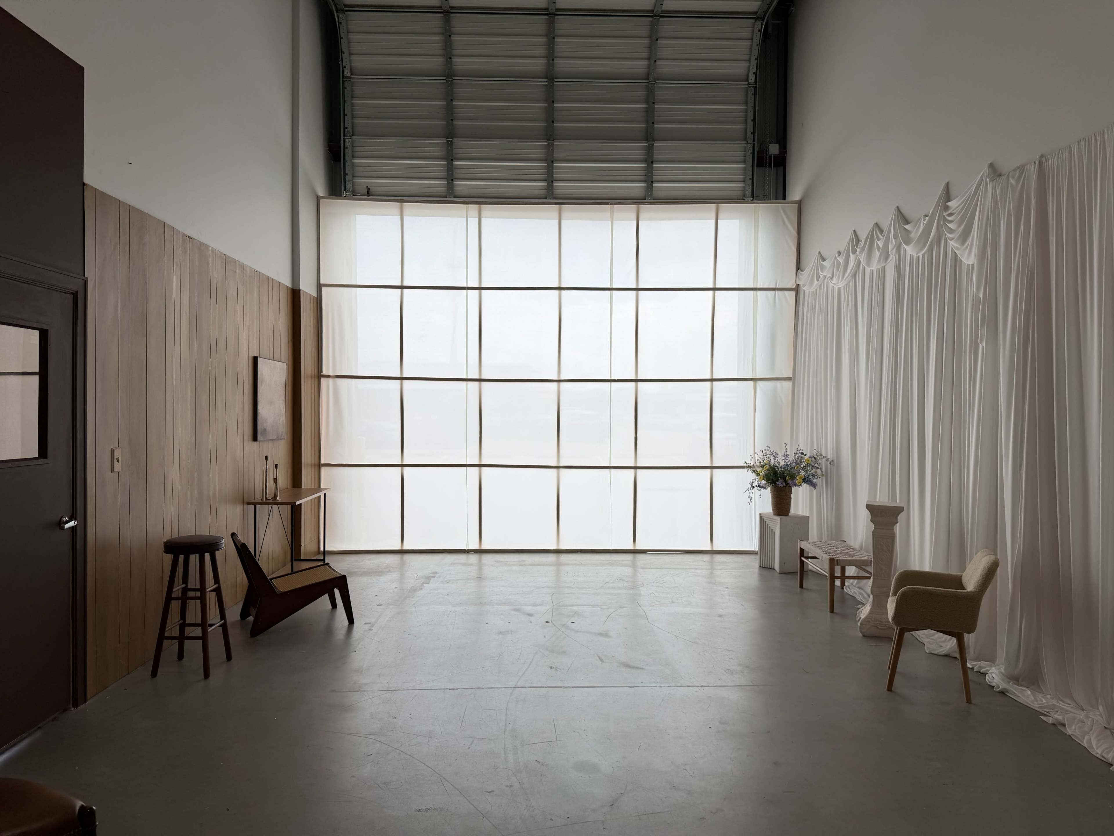 The image depicts an empty interior space with a large window lined with sheer curtains, accompanied by minimalist furniture including a chair and a small table.