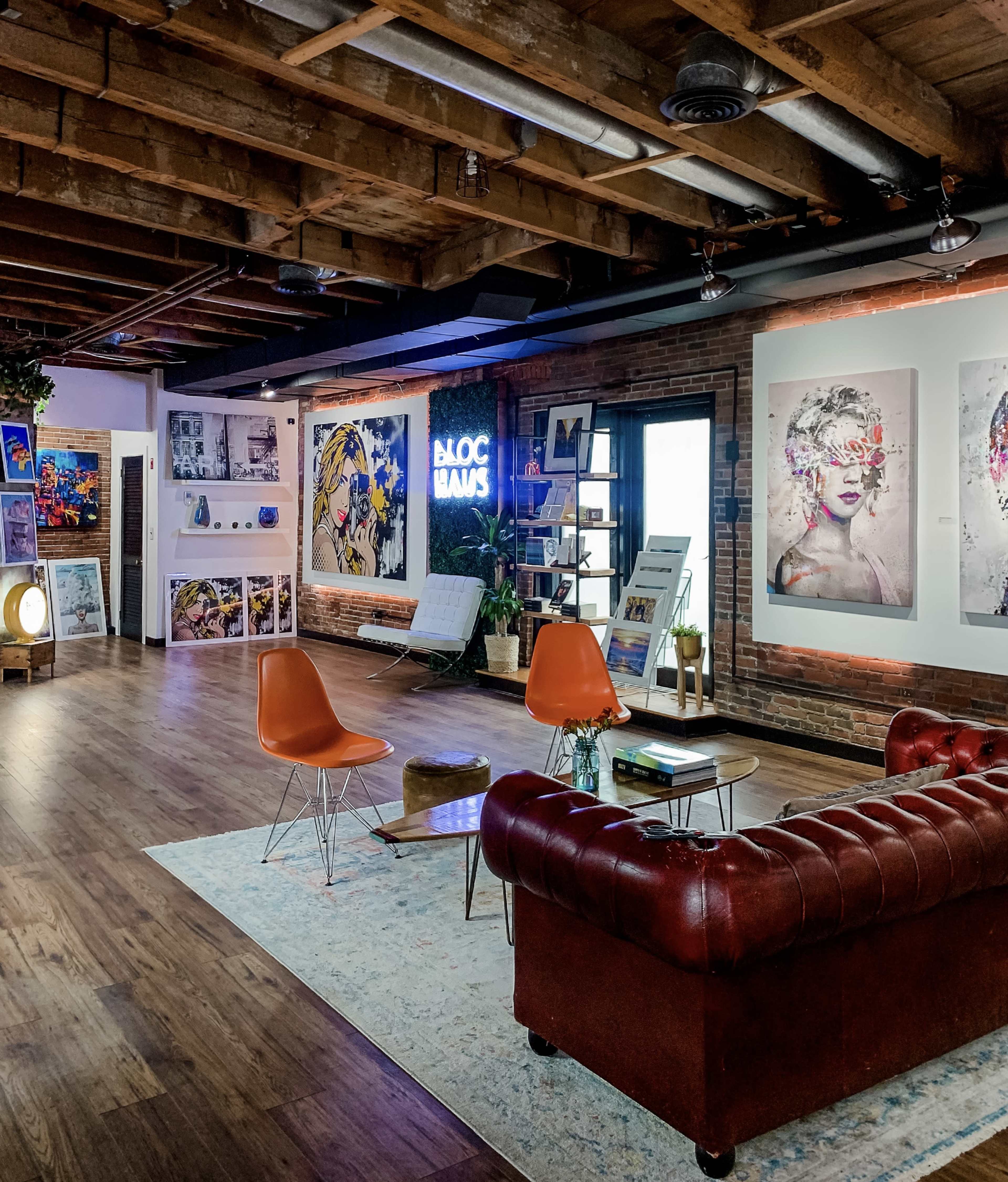 A contemporary art studio featuring a mix of colorful artwork on the walls, a red leather sofa, orange chairs, and wooden beams on the ceiling.