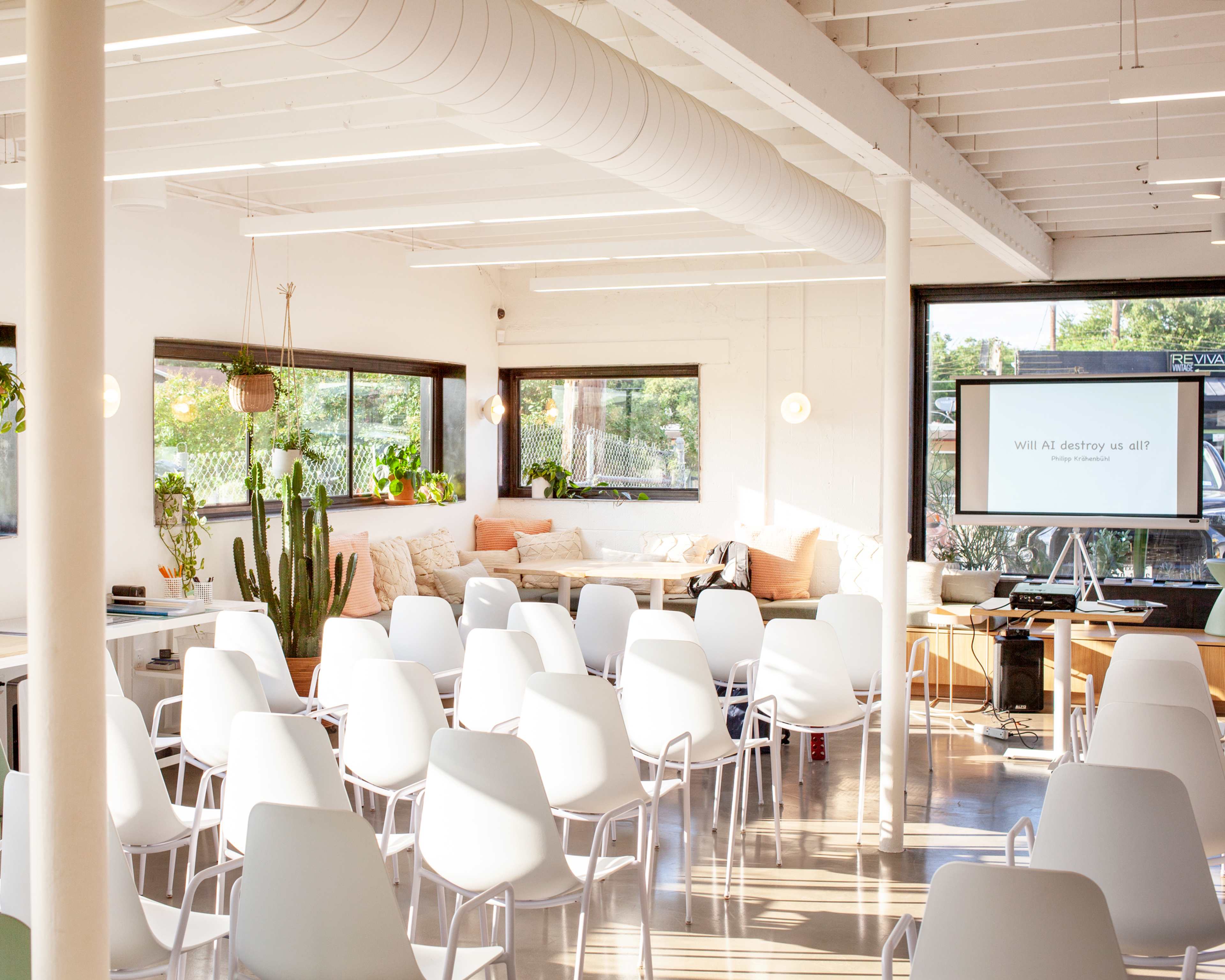 A bright, open meeting space features rows of white chairs facing a screen, with large windows allowing natural light and plants decorating the area.