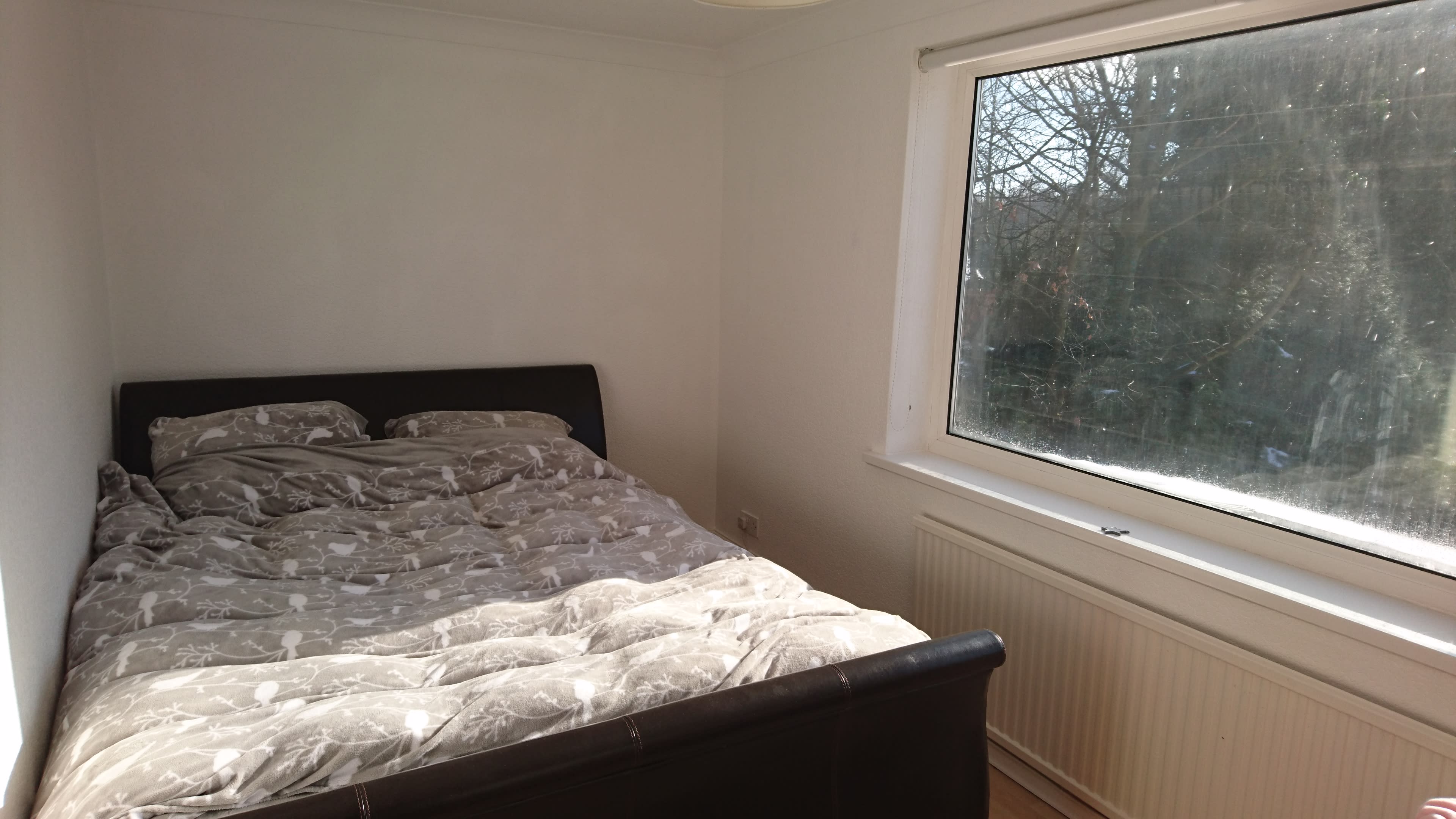 A light-filled bedroom features a double bed with gray patterned bedding against a white wall and a large window overlooking a garden.