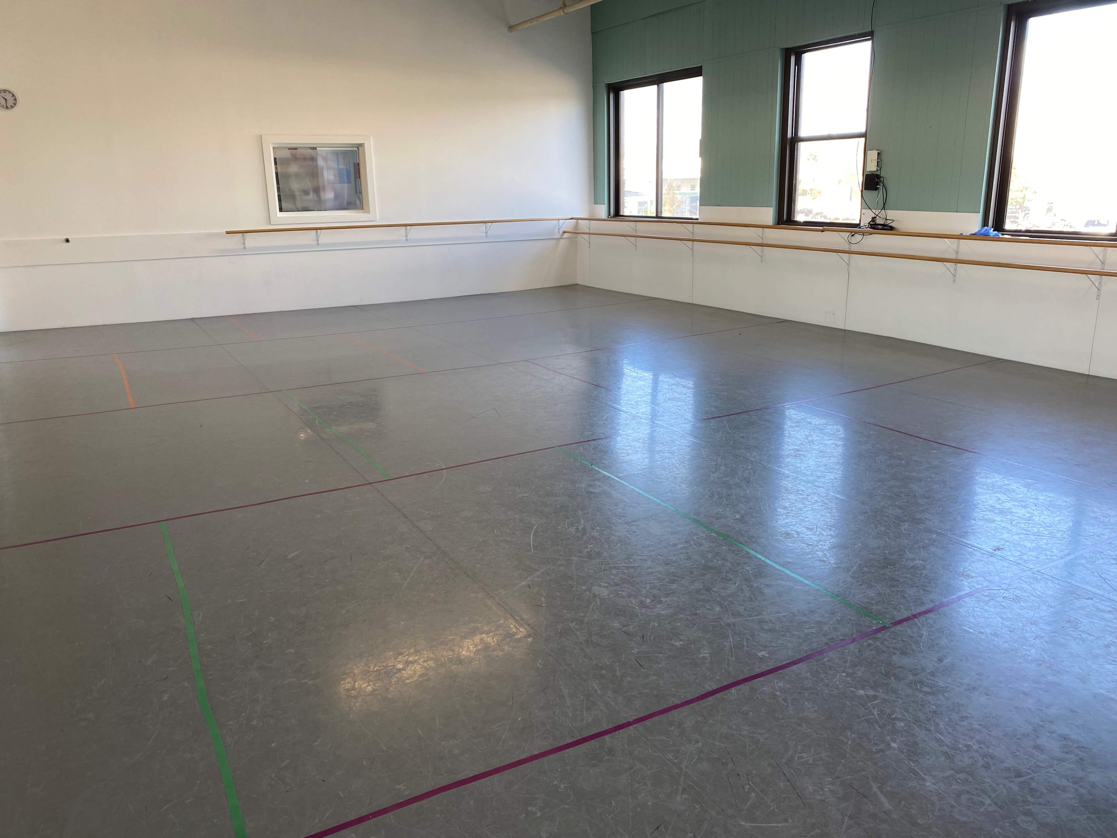 A spacious dance studio with a smooth, grey floor, marked by colored lines, and large windows providing natural light.