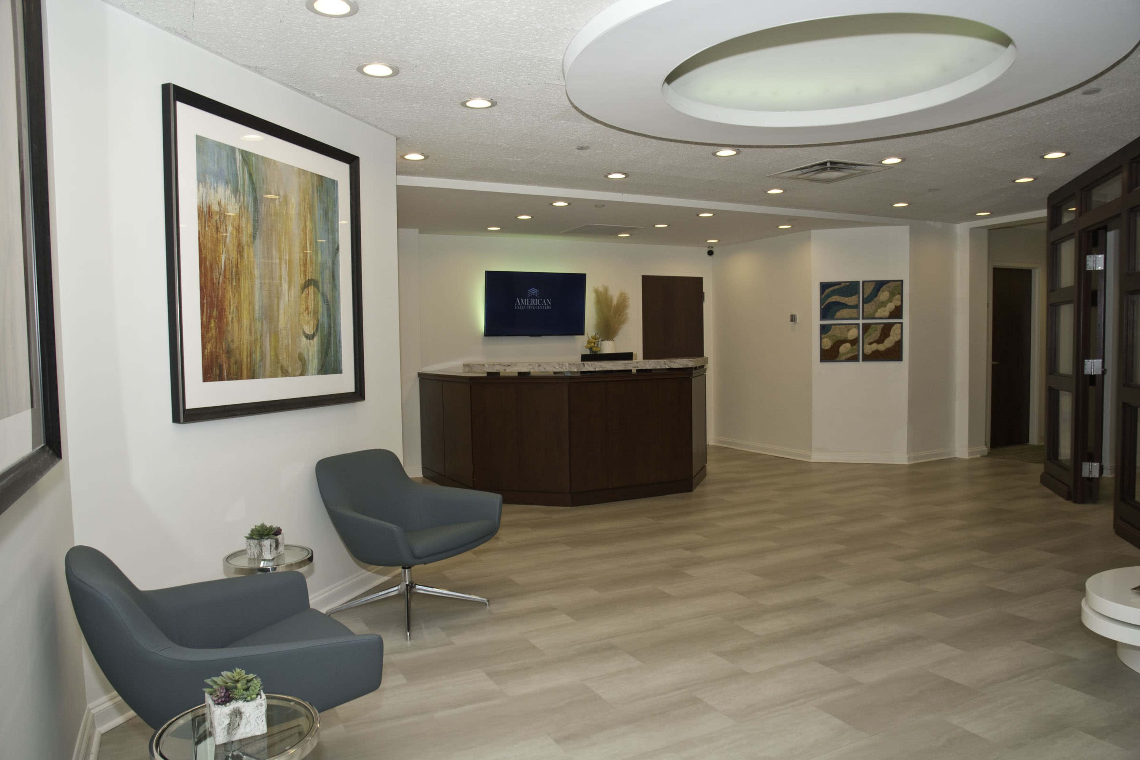 The image shows a modern reception area with two gray chairs, a front desk, and artwork hanging on the walls.