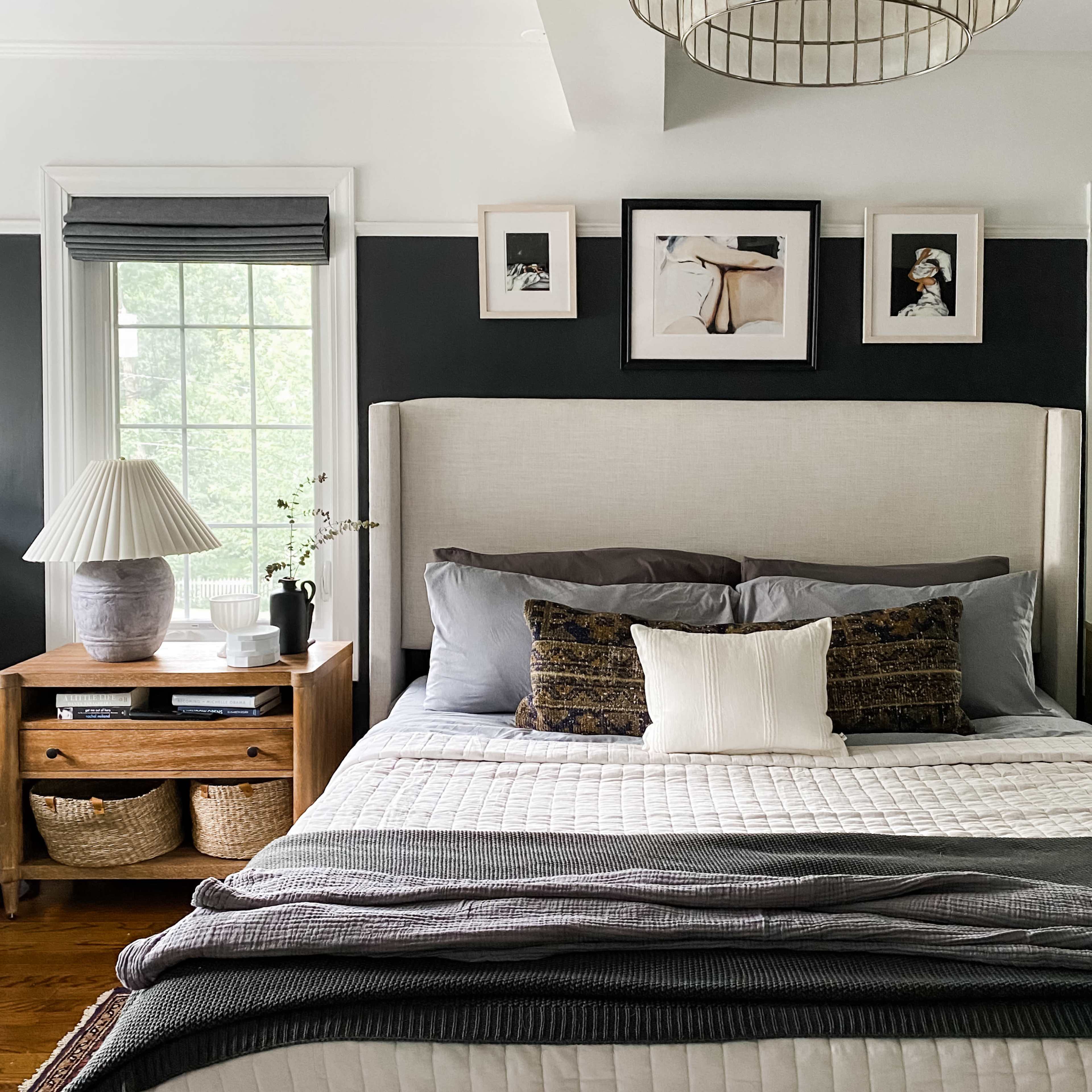 A well-decorated bedroom featuring a light-colored upholstered headboard, a bed with layered gray and white bedding, and a bedside table with decorative items and storage baskets.