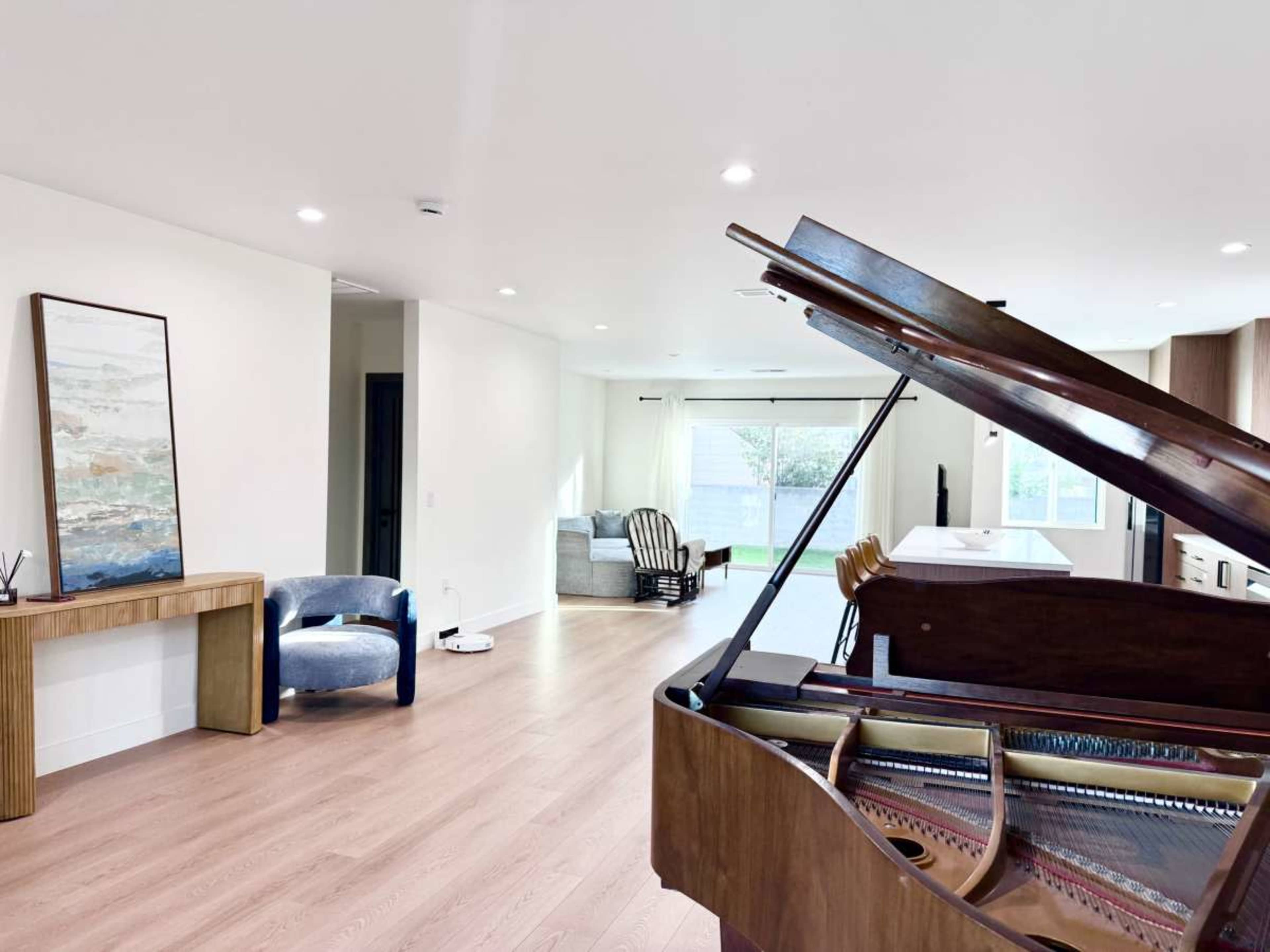 The image shows a spacious room with a grand piano in the foreground, a wooden console table, a blue accent chair, and a gray couch in the background with large windows.