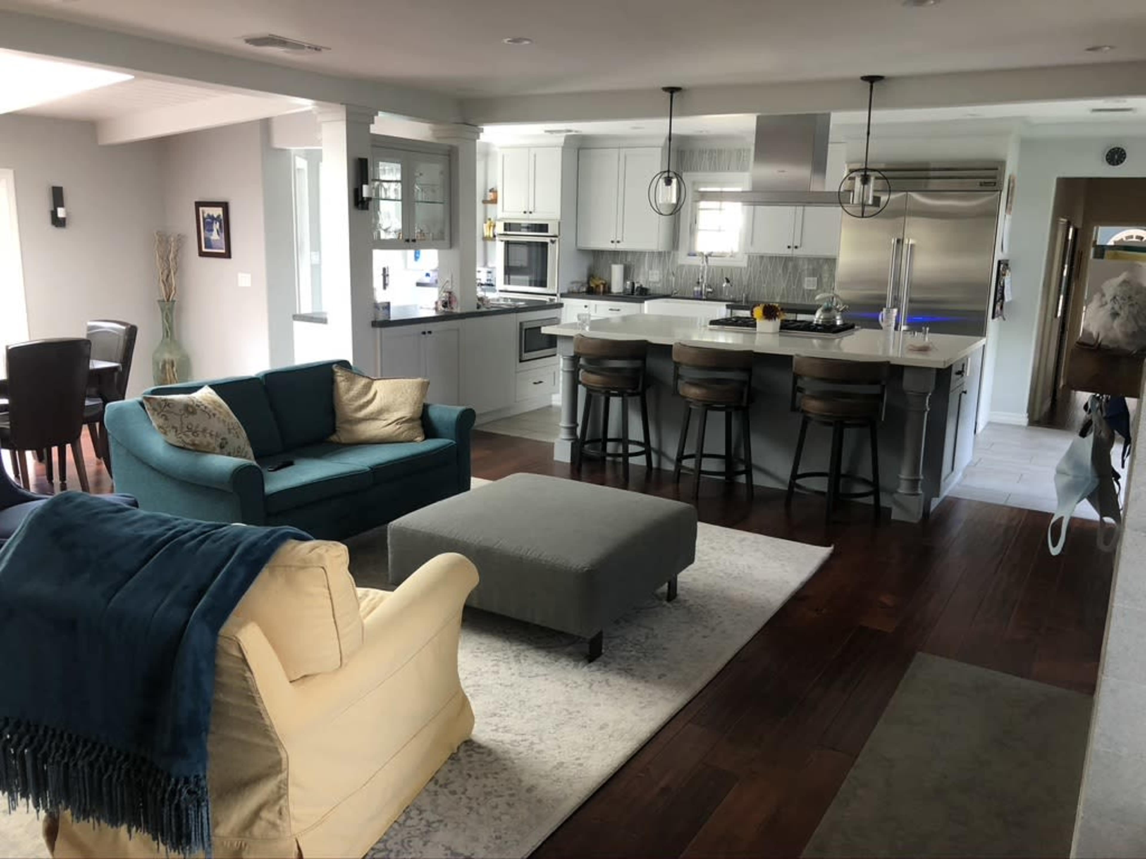 A modern open-concept living room and kitchen area with a blue couch, beige chair, and a gray ottoman on a light-colored rug.