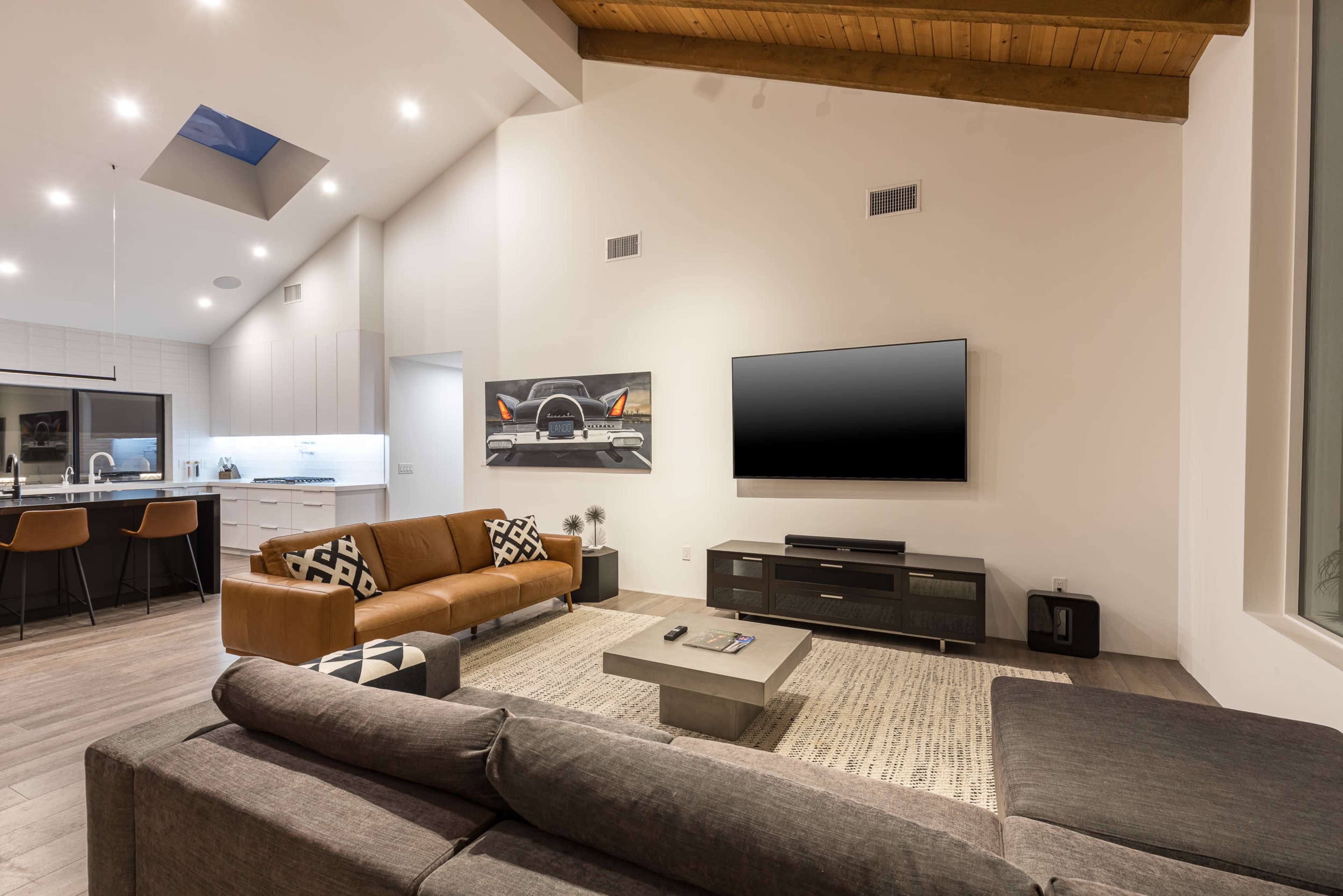 A modern living room features a large TV mounted on a light-colored wall, a brown leather sofa, a gray sectional couch, and an open view of a kitchen with bar stools in the background.