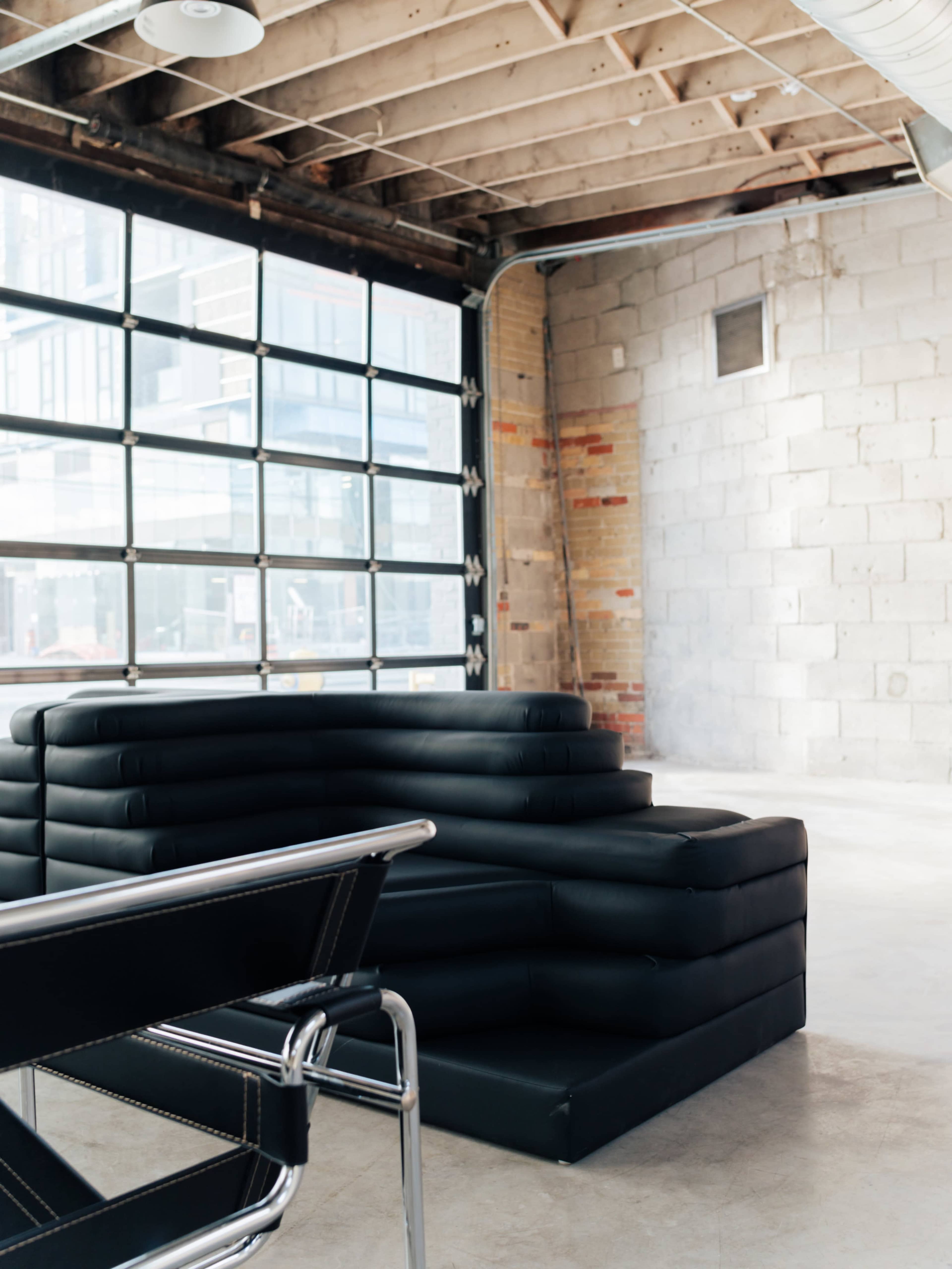 A black modular sofa is positioned in a spacious industrial-style room with large windows and exposed brick walls.
