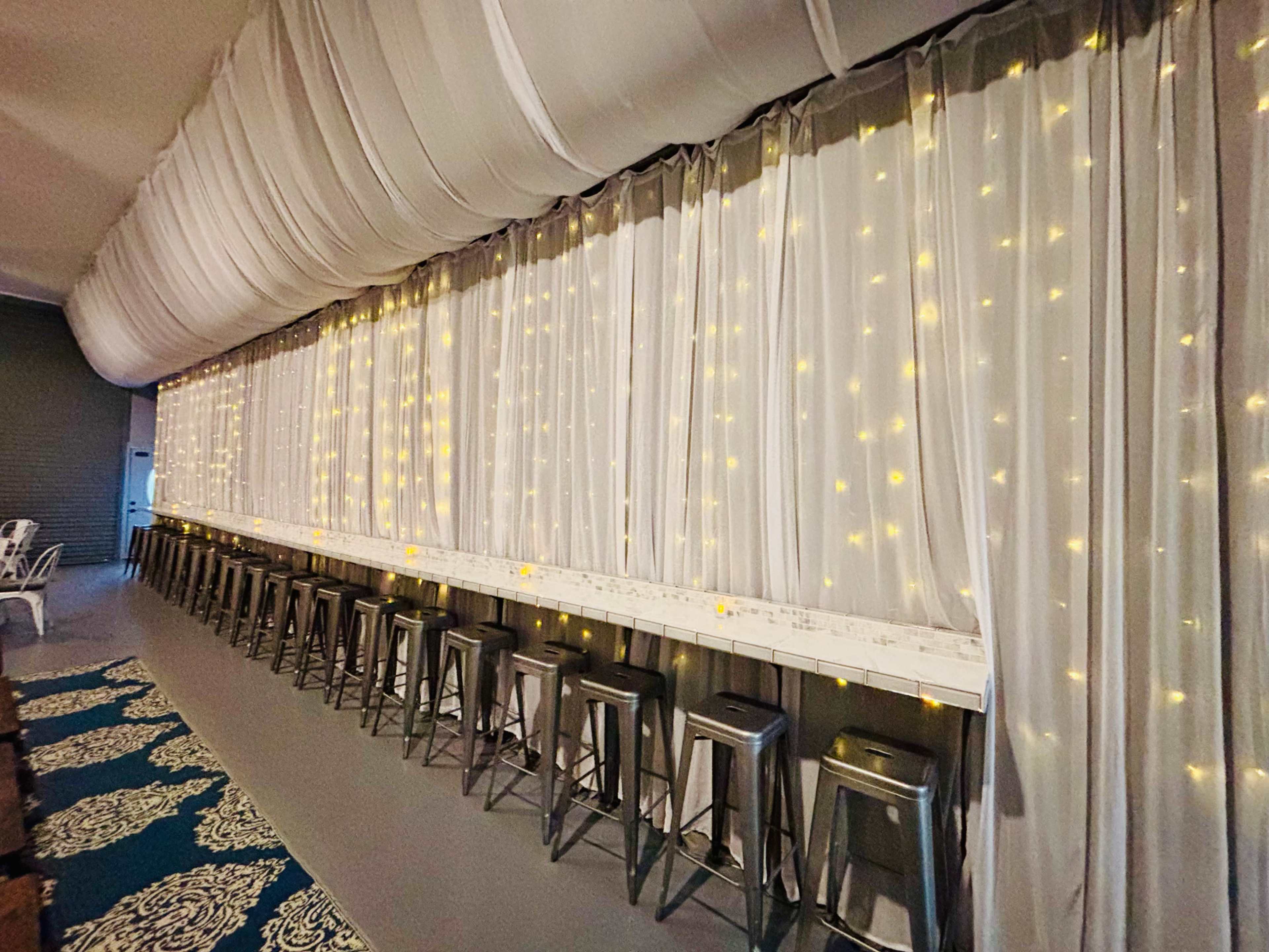 A row of metal stools is lined up against a wall covered with white curtains and ambient string lights.