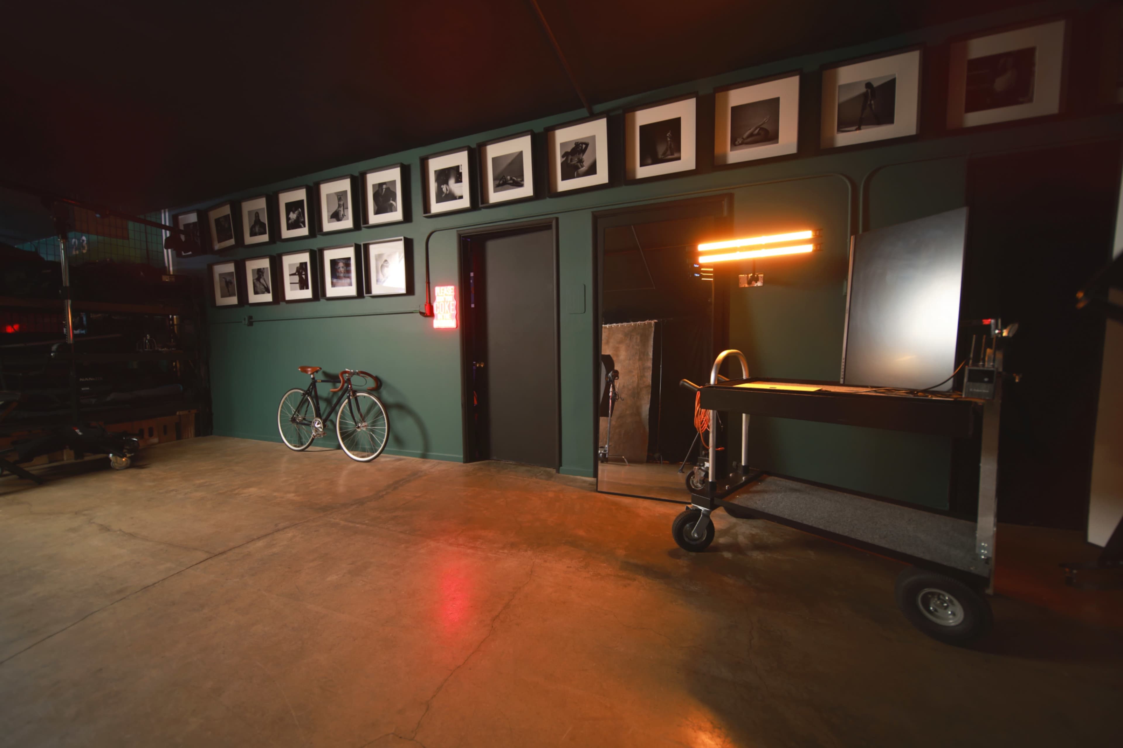The image shows a dimly lit room with a green wall adorned with black-and-white photographs, a bicycle leaning against the wall, and a cart with a light source positioned nearby.