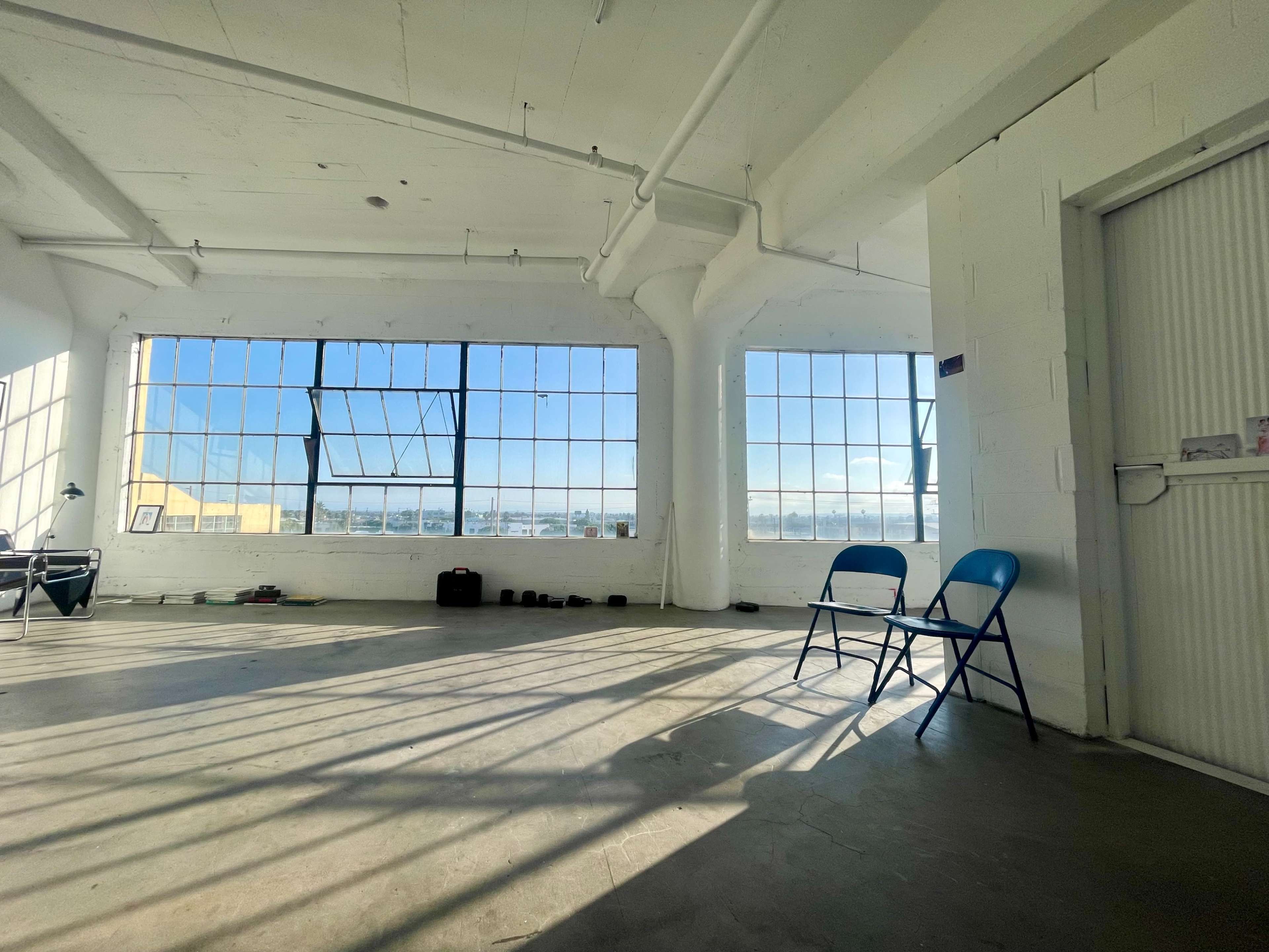 A spacious room with large windows providing sunlight, two blue chairs, and some weights on the floor.