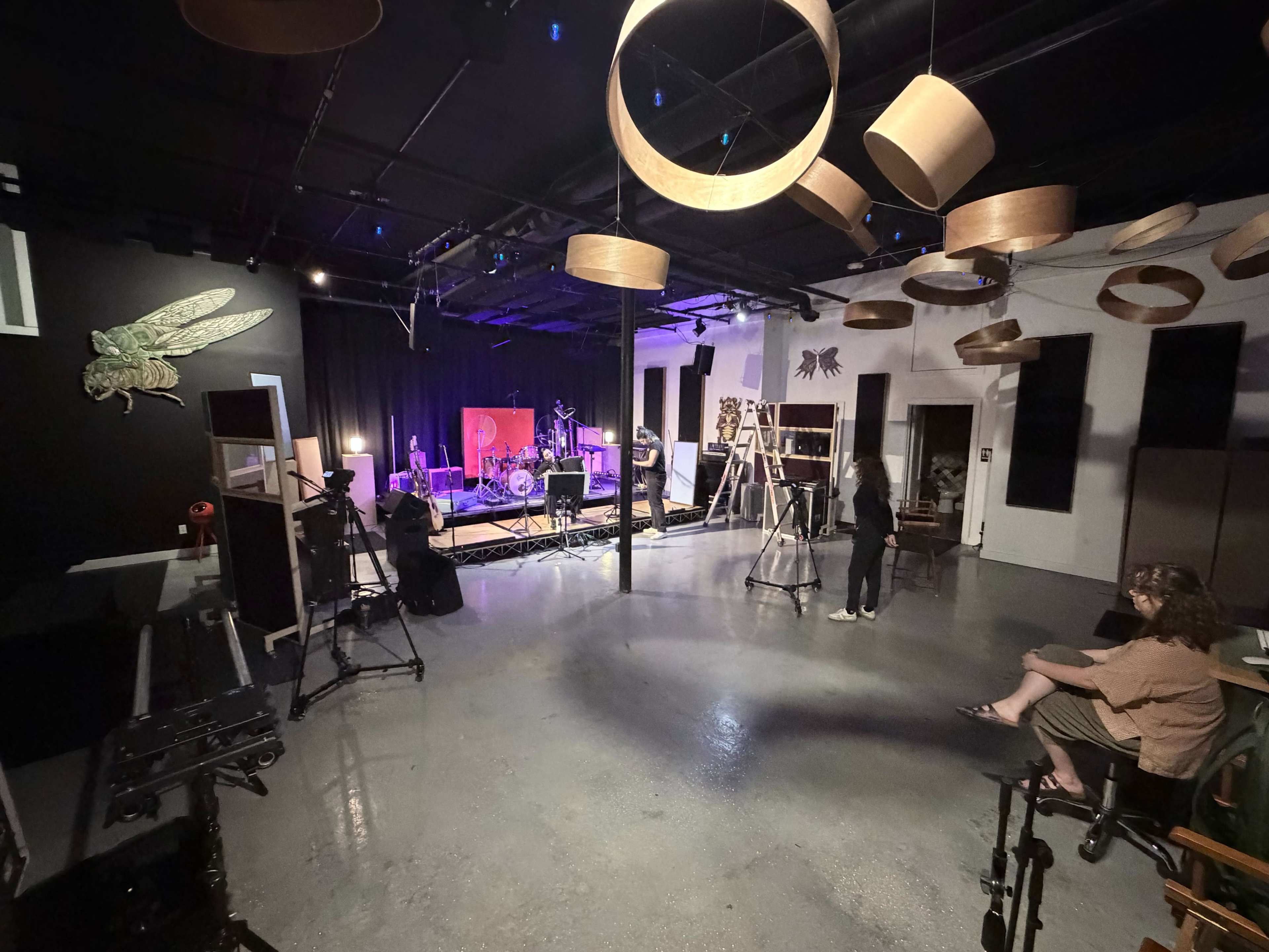 The image shows a spacious, dimly lit studio with a small stage set up for a live performance, featuring musical instruments and equipment, while individuals are positioned throughout the room.