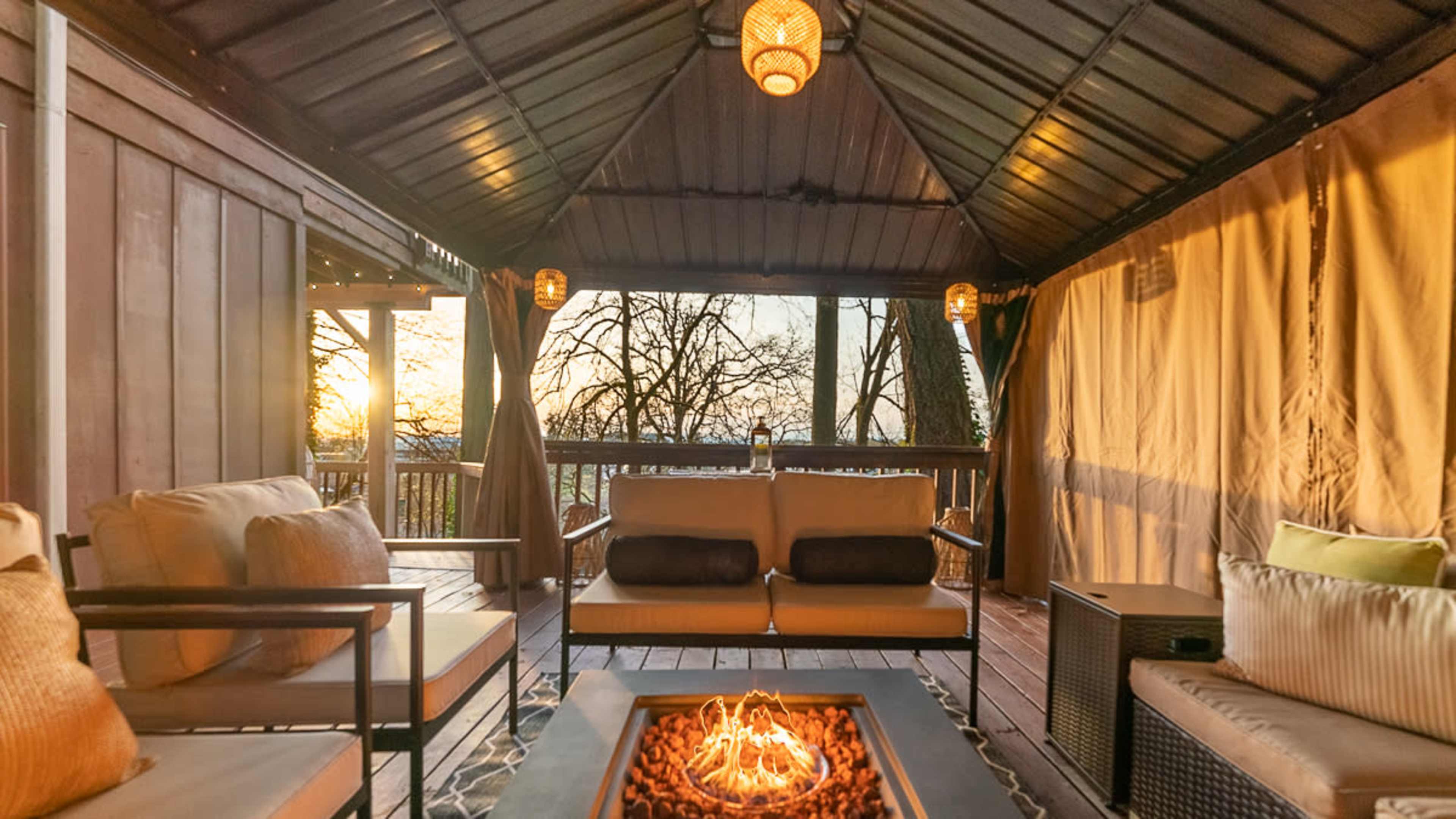 A cozy outdoor seating area with a fire pit, surrounded by draped curtains and overlooking a sunset through trees.