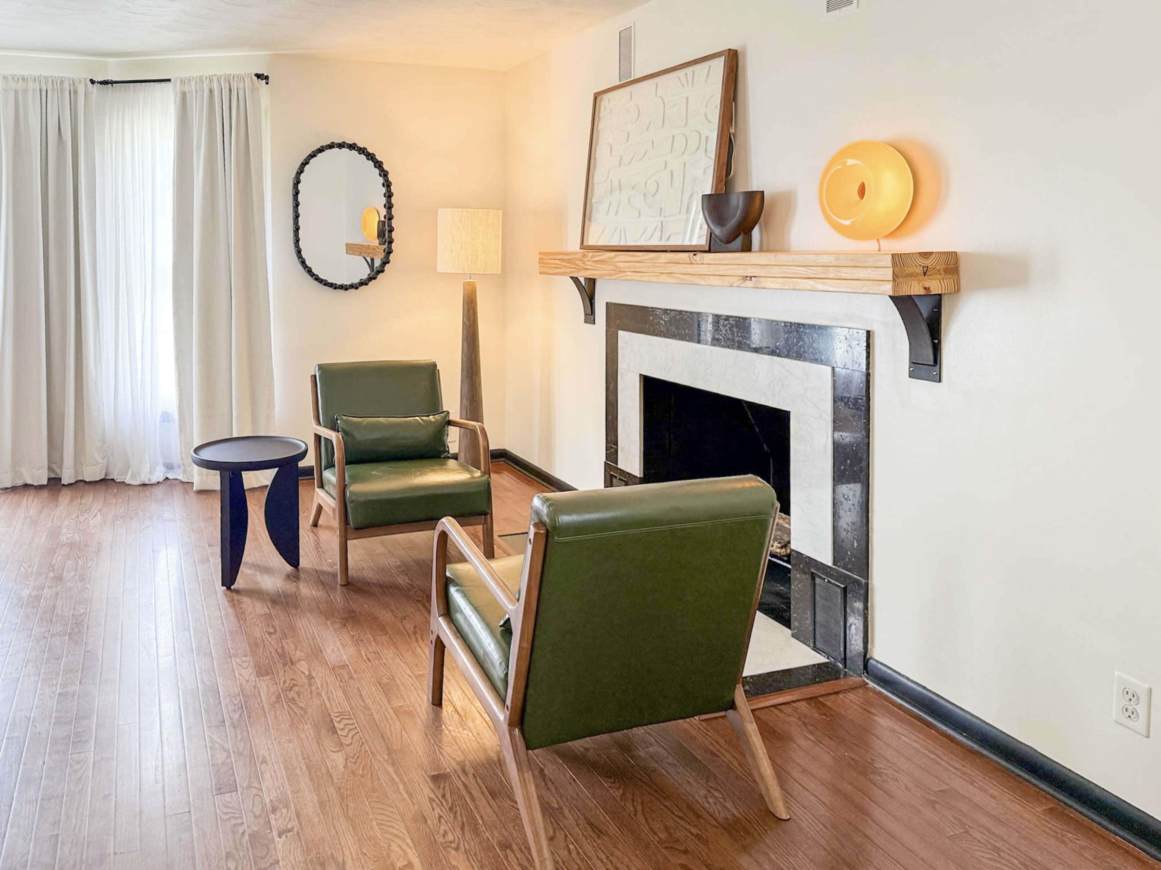 The image shows a living room featuring two green armchairs, a small black side table, a modern lamp, and a fireplace with decorative items on a wooden shelf.