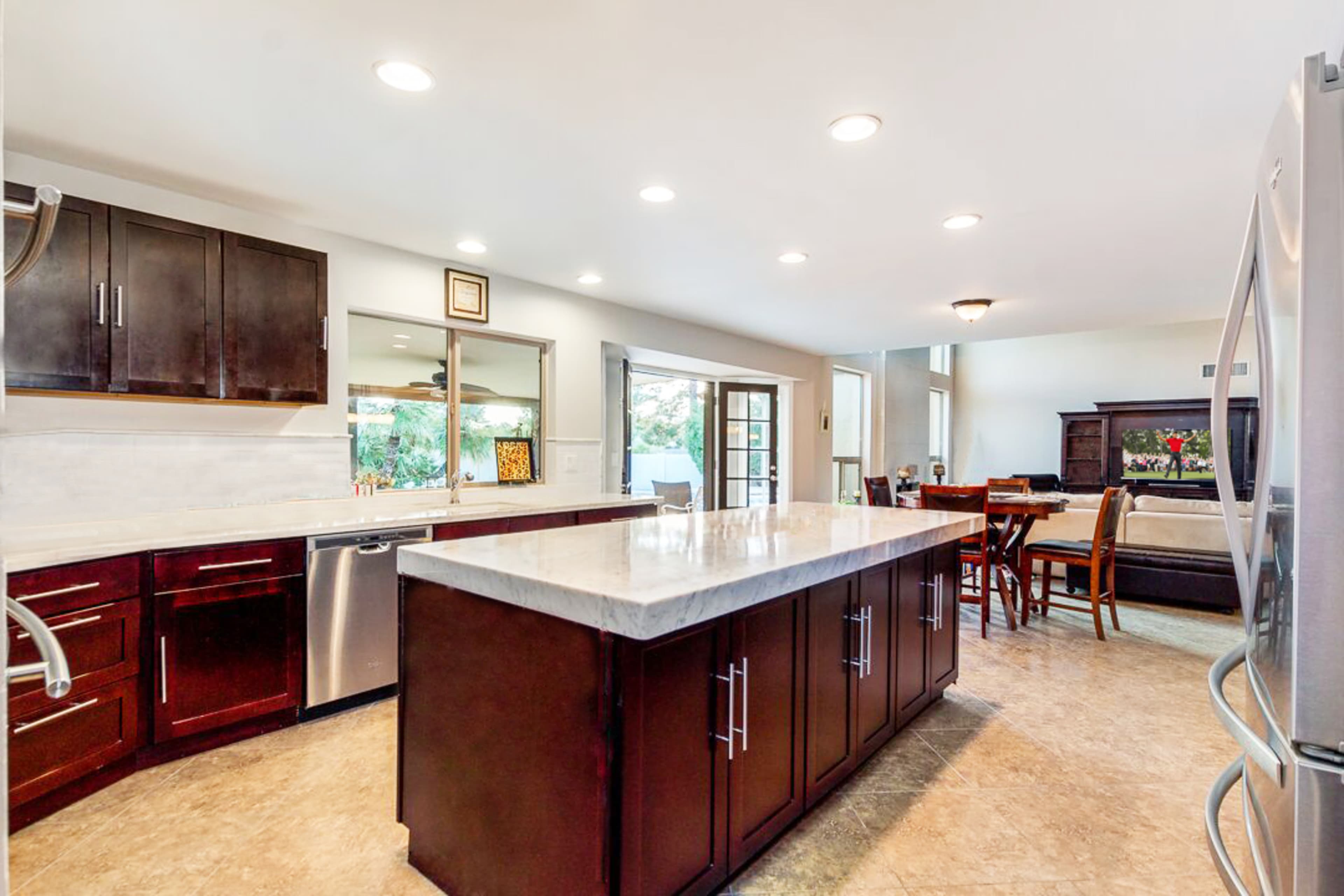 A modern kitchen with dark wooden cabinets, a marble countertop island, and large windows allowing natural light to enter.