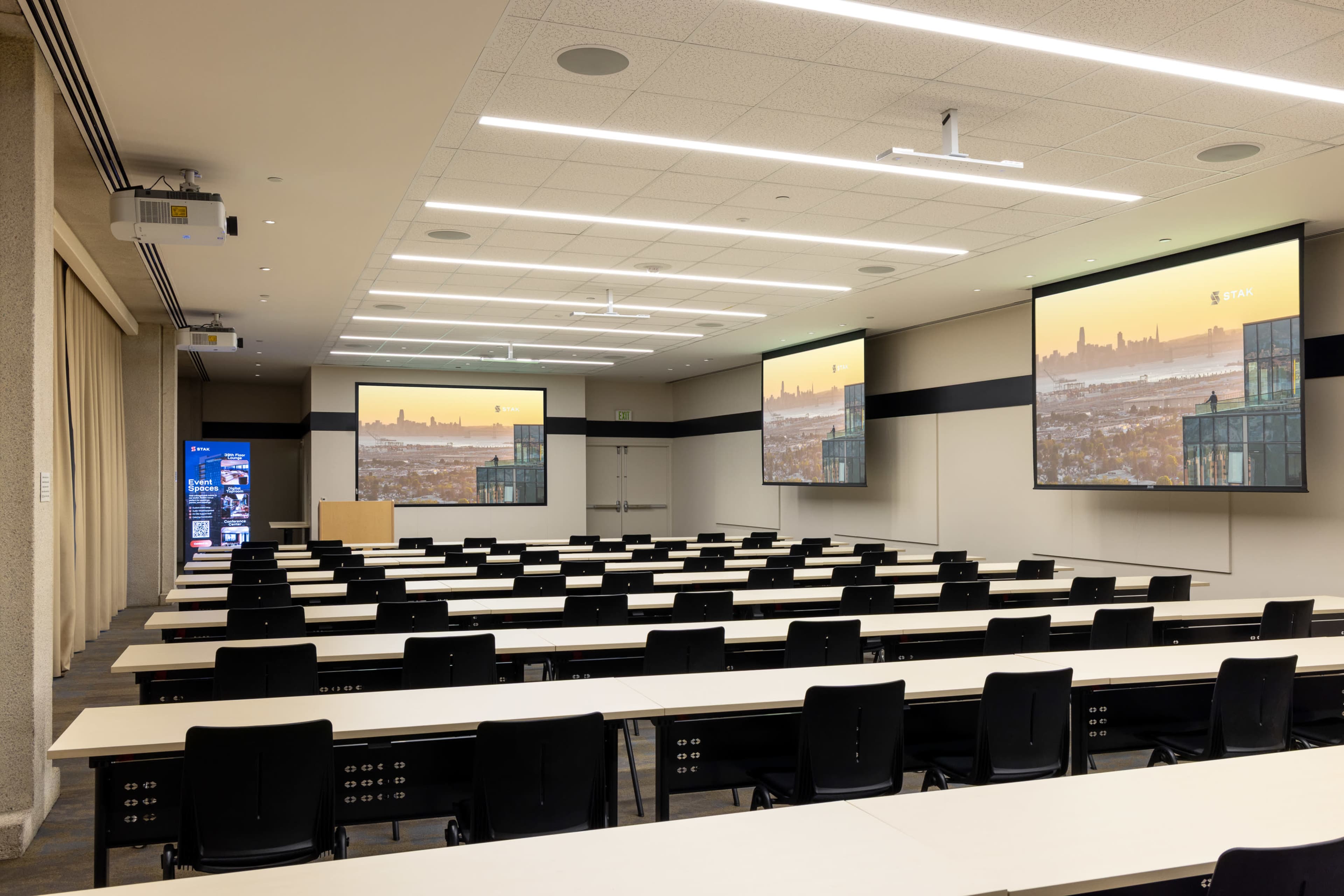 A modern conference room features multiple rows of tables and chairs arranged in front of large screens displaying a city skyline.