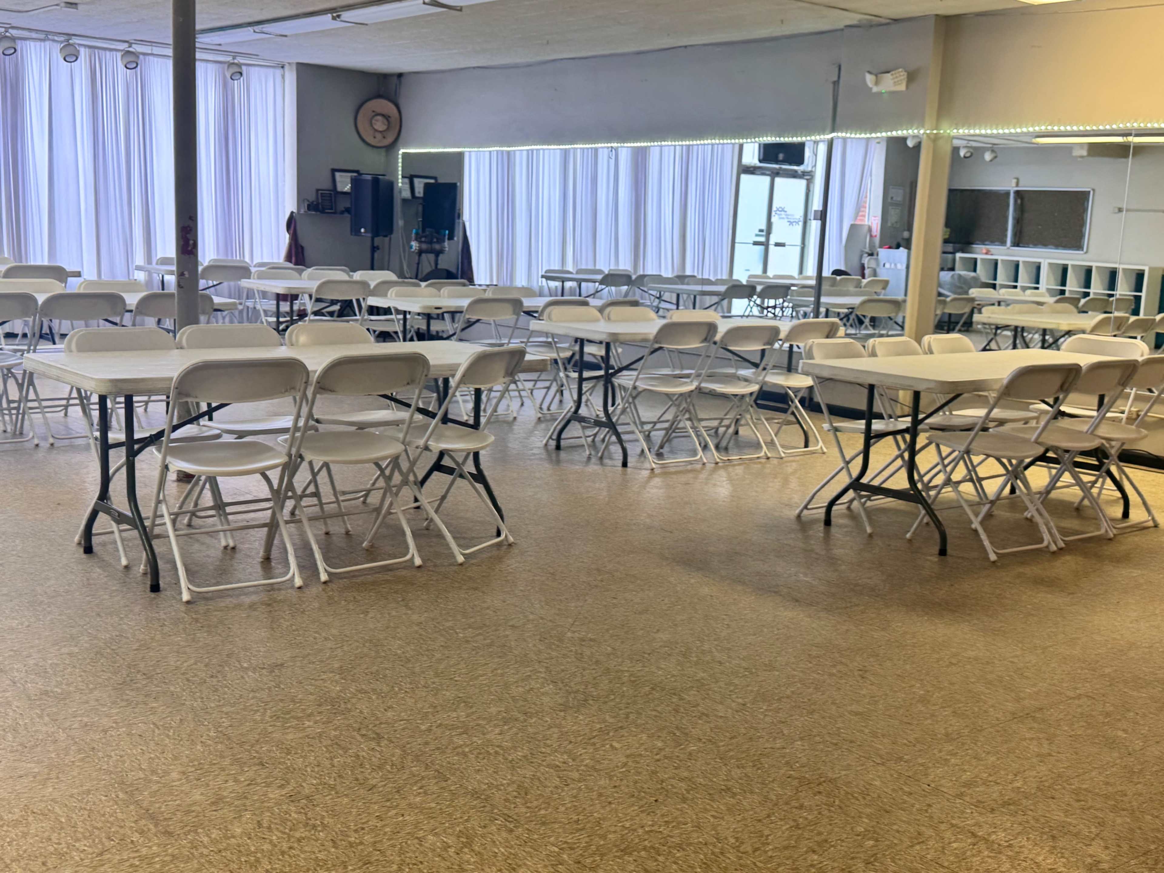 The image shows a spacious room with white folding chairs arranged around several tables, featuring large windows with curtains that allow natural light to enter.