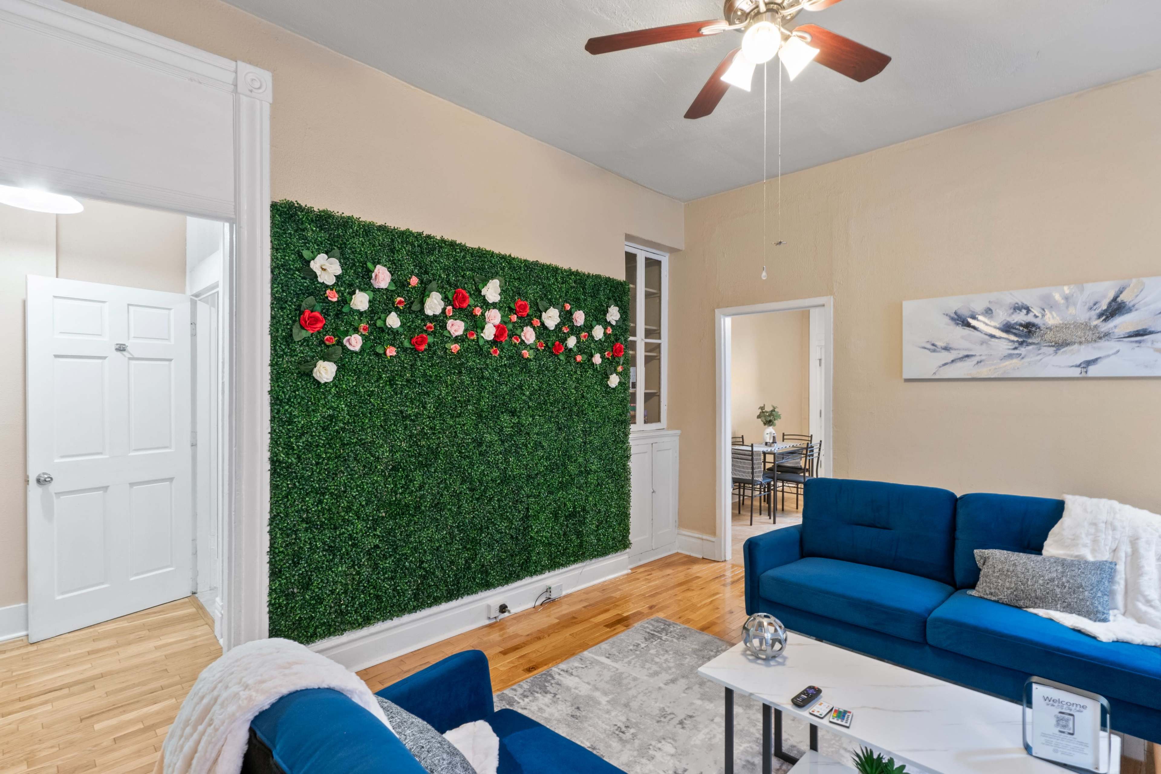 The living room features a green wall adorned with artificial flowers, flanked by two blue sofas and a light-colored rug.