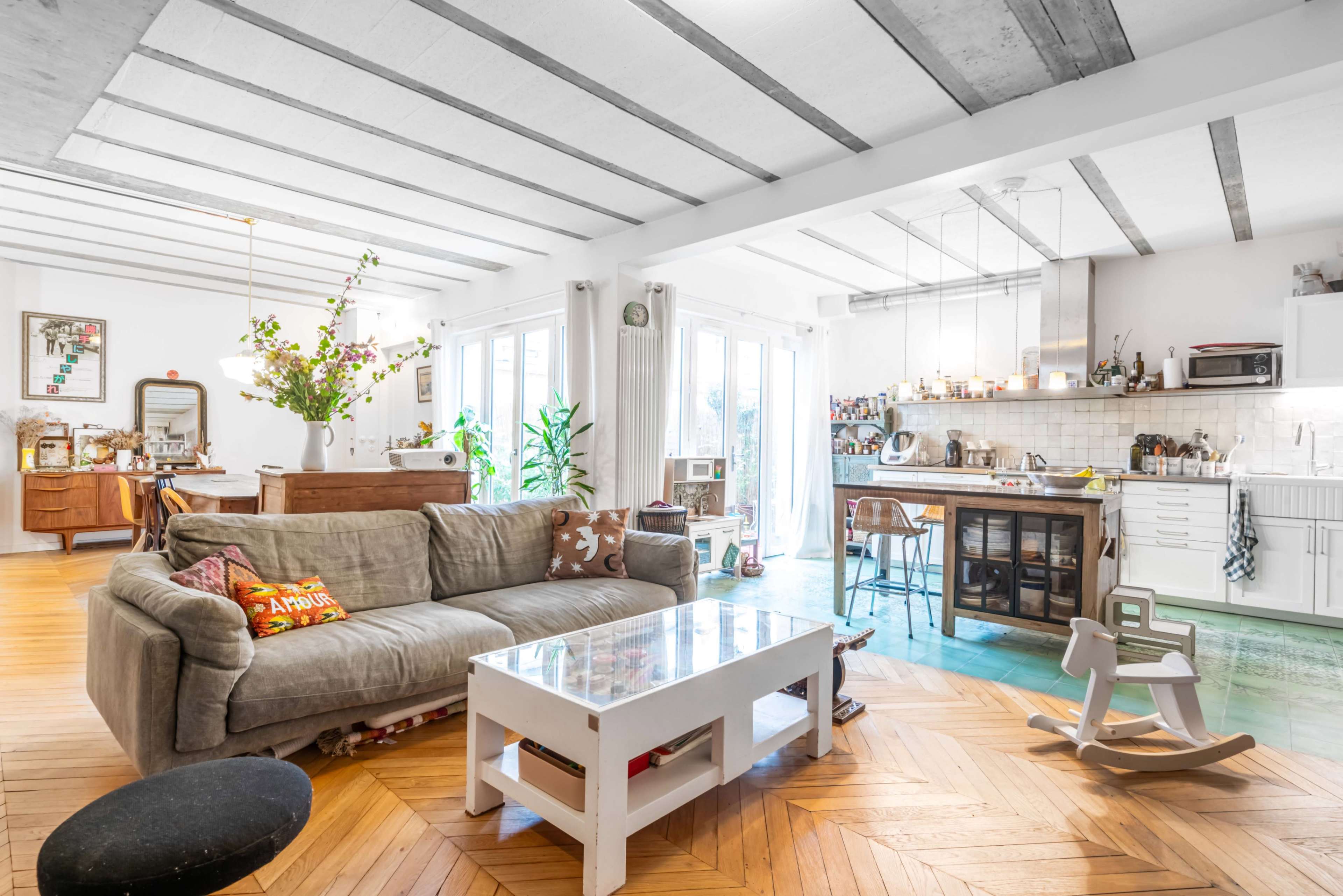 The image shows a spacious, well-lit living area featuring a gray sofa, a glass coffee table, and a kitchen in the background with a mix of modern and vintage decor.