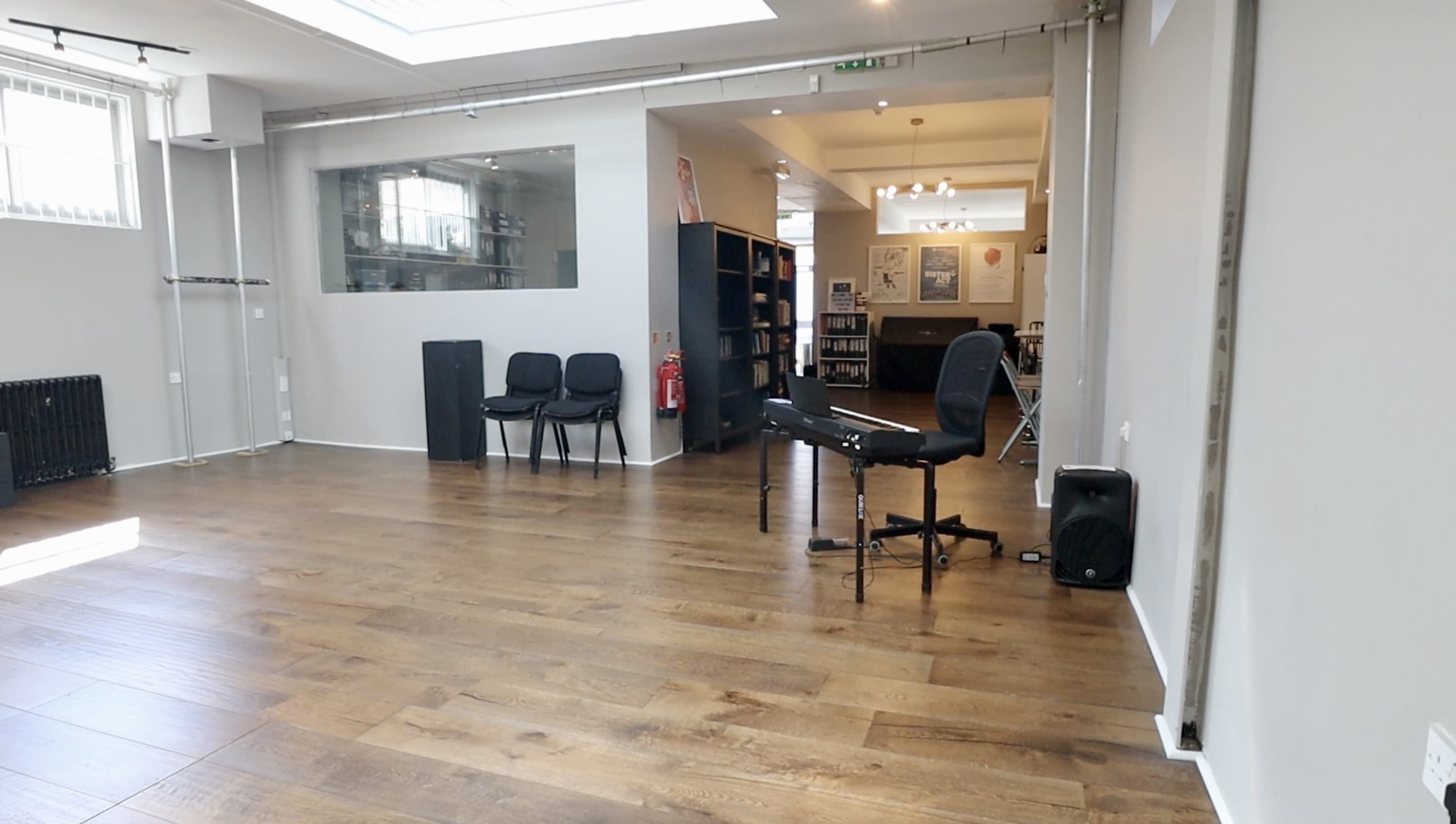 A spacious, empty room with wooden flooring, featuring a few chairs, a black office chair, and a desk, along with shelves and a windowed area in the background.